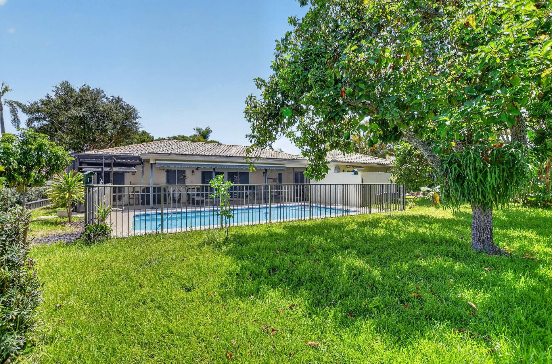 Fenced swimming pool, paved patio area, rear exterior with multiple windows, mature landscaping, and a tiled roof