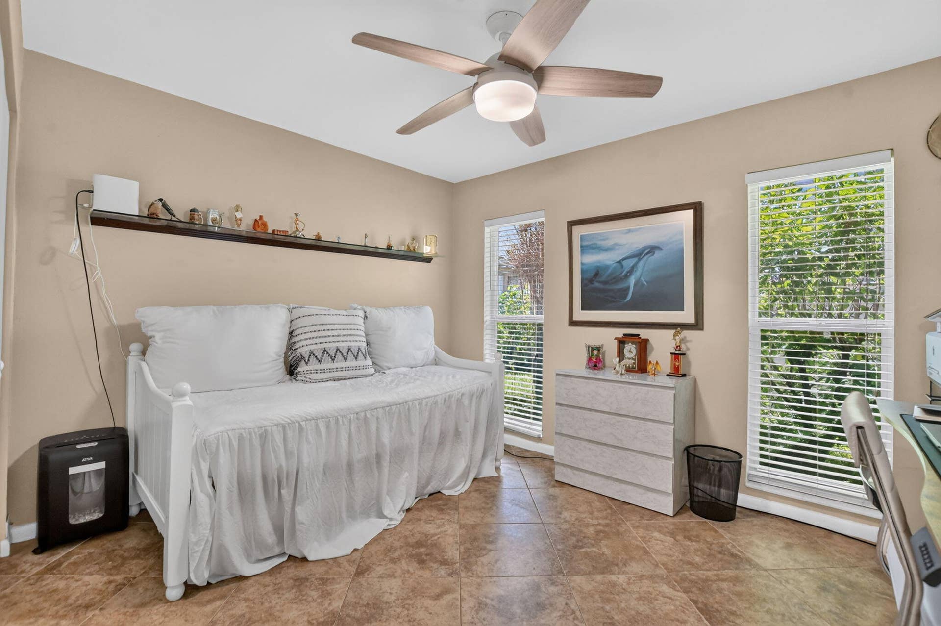 Neutral wall color, ceiling fan with integrated lighting, two windows with blinds, built-in wall shelving, and tile flooring