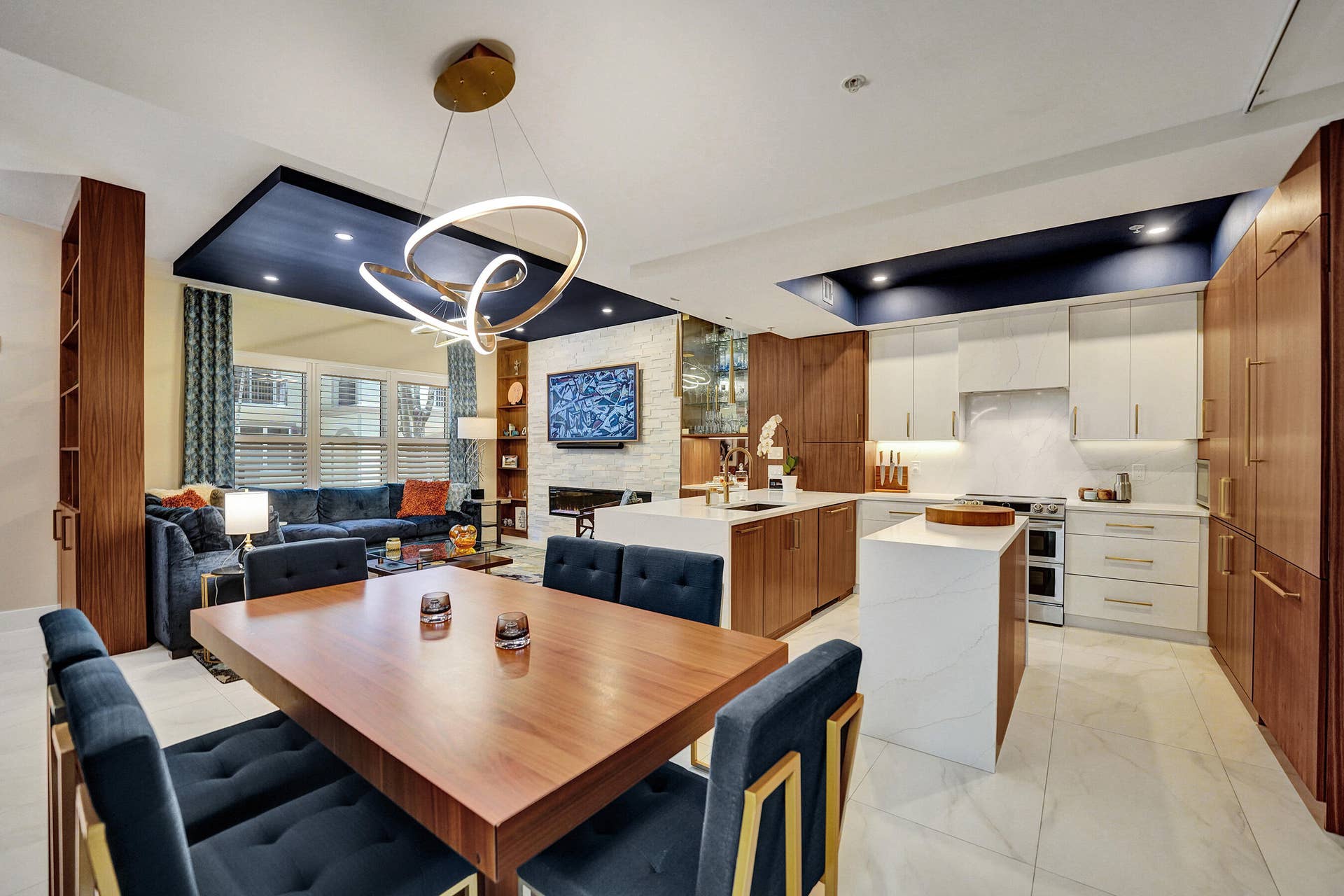 Open-concept living area with a contemporary chandelier, built-in shelving, a kitchen island with a waterfall countertop, integrated paneled appliances, and a stacked stone fireplace