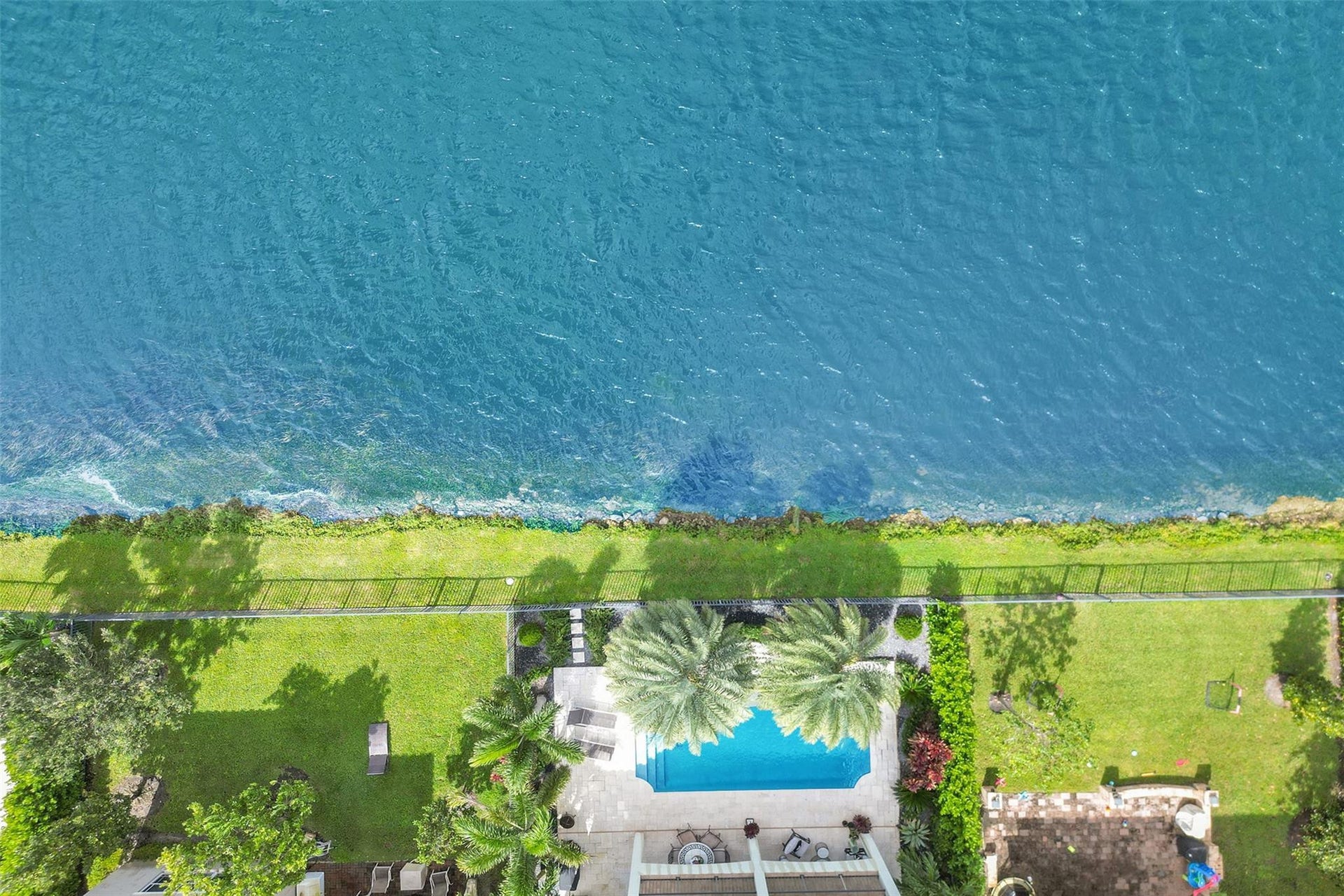 9749 Blue Isle, #BAY, Parkland, FL 33076 Waterfront property with a rectangular swimming pool, expansive lawn, mature palm trees, and a paved pool deck