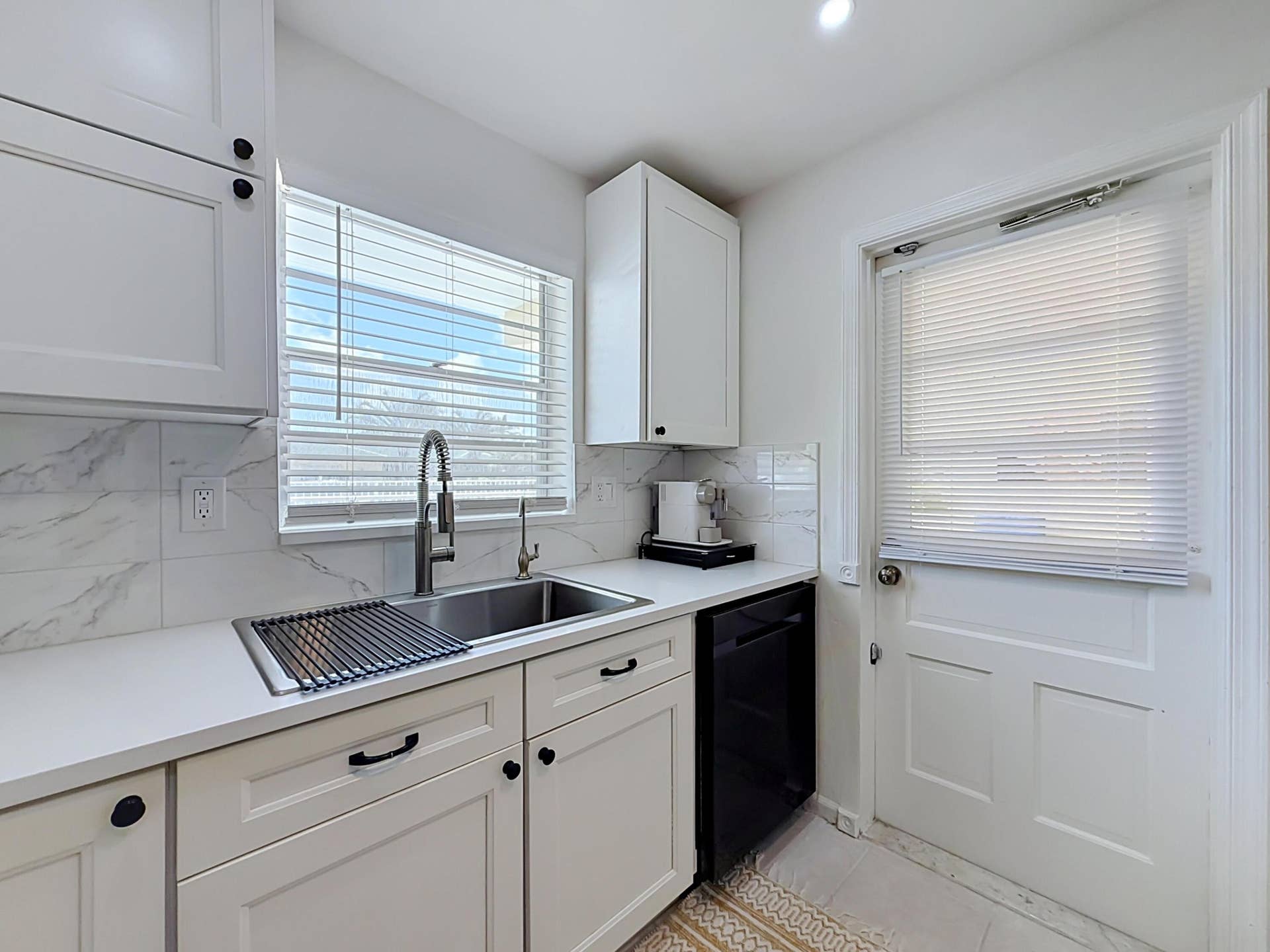White shaker-style cabinetry with black hardware, undermount stainless steel sink, pull-down faucet, white solid surface countertops, and marble-look tile backsplash