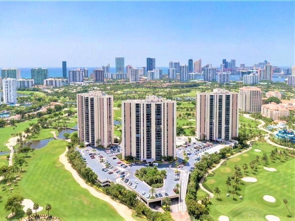 20335 W COUNTRY CLUB Drive, #2408, Aventura, FL 33180 Three high-rise buildings situated amidst a golf course landscape with a distant city skyline and ocean background