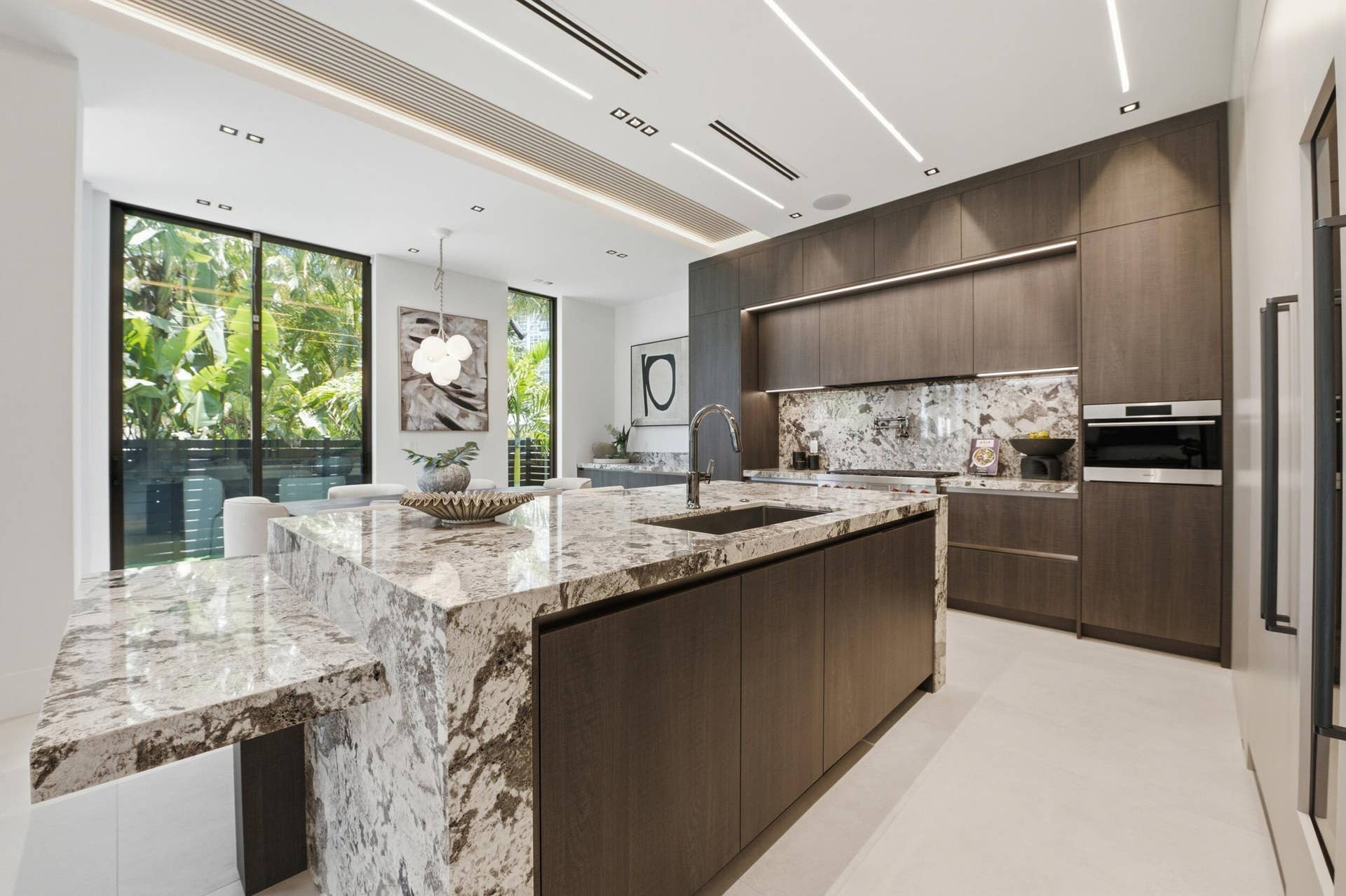 Spacious kitchen featuring a large island with an integrated breakfast bar, dark wood-finish cabinetry, built-in oven, and a complementary stone backsplash