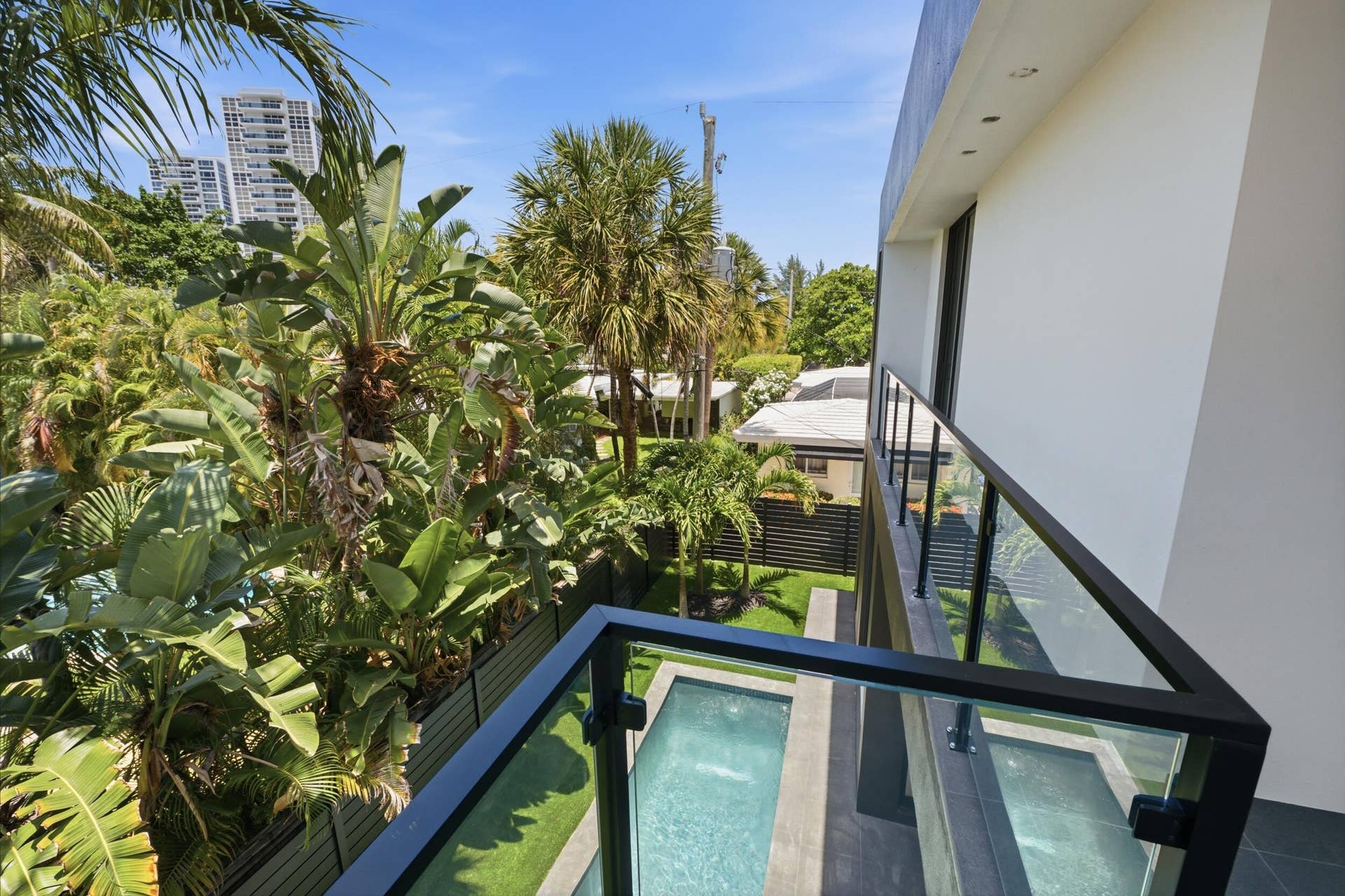 Balcony with glass panel railing and black frame, overlooking a private pool, artificial turf, and lush tropical landscaping