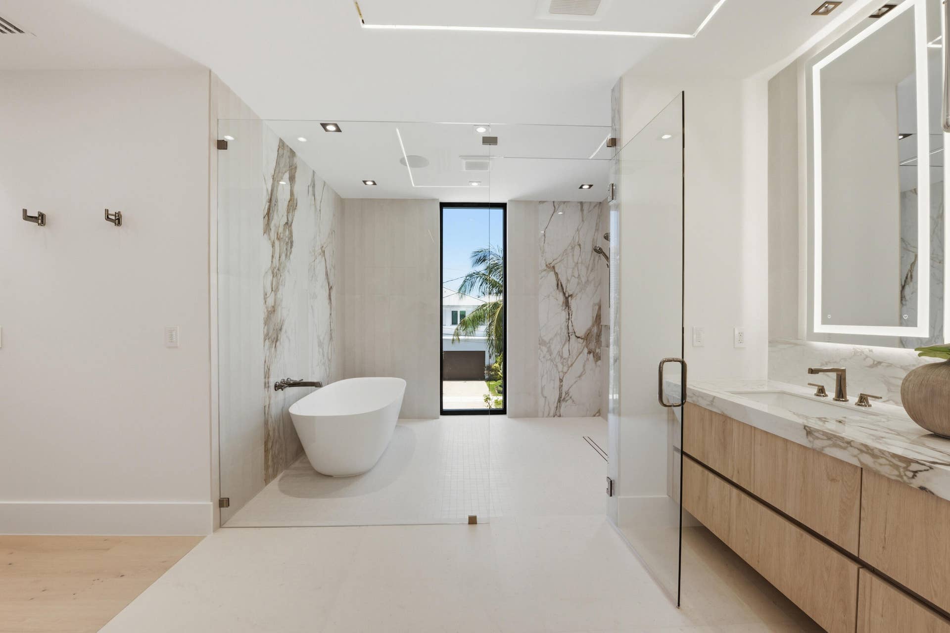 Freestanding soaking tub, floor-to-ceiling window, wood-finish vanity with stone countertop, backlit mirror, and glass-enclosed shower