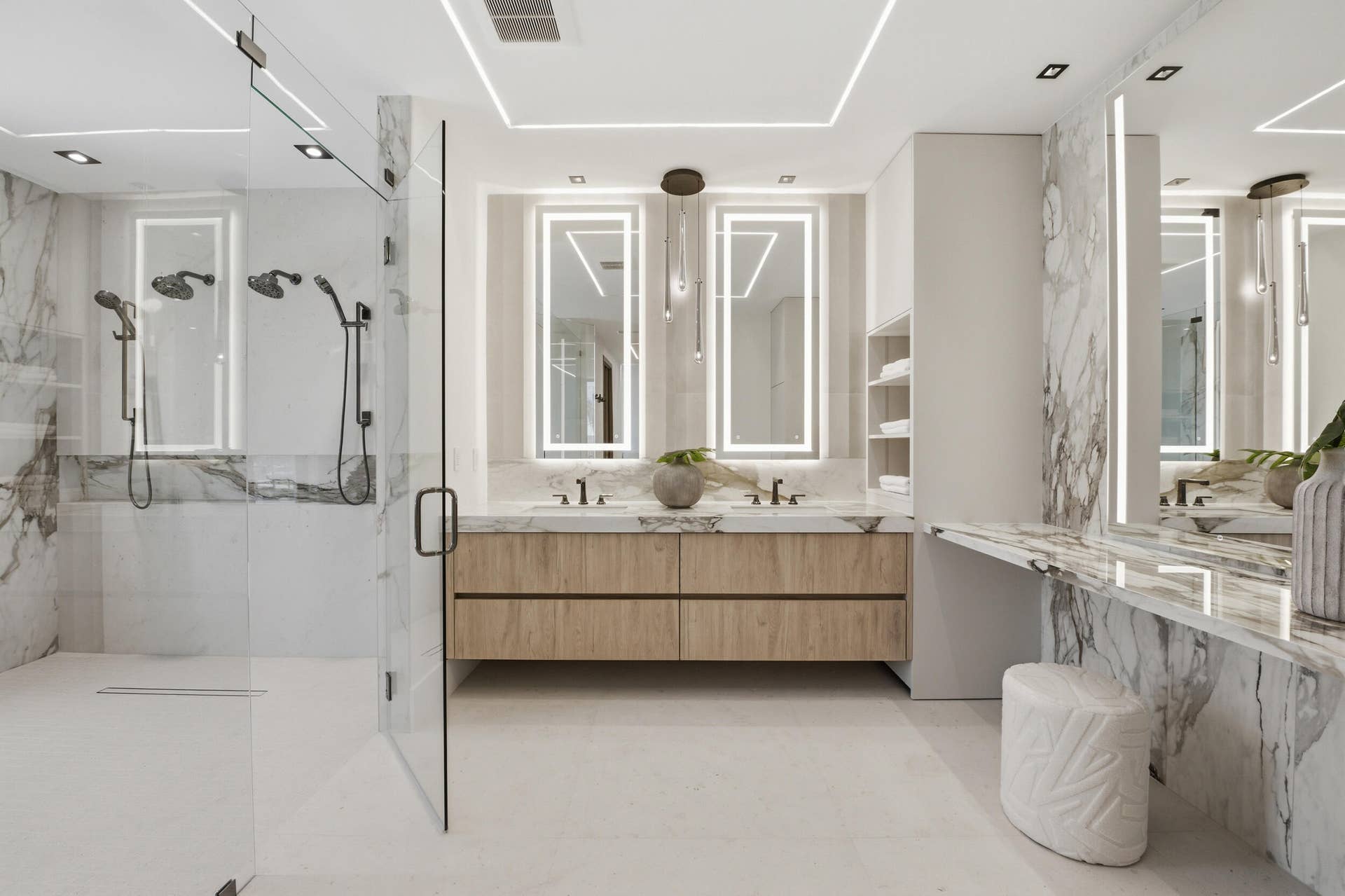 Glass-enclosed shower with multiple showerheads, dual floating vanities with integrated lighting, marble-patterned wall and vanity surfaces, and recessed ceiling lighting