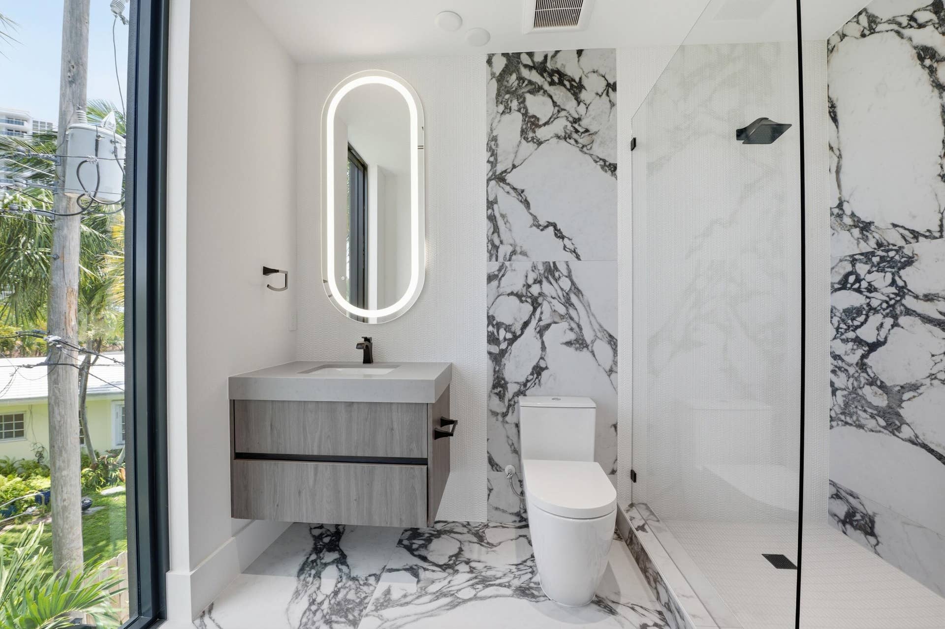 Floating vanity with integrated sink, backlit oval mirror, floor-to-ceiling tile, glass-enclosed shower, and full-height window
