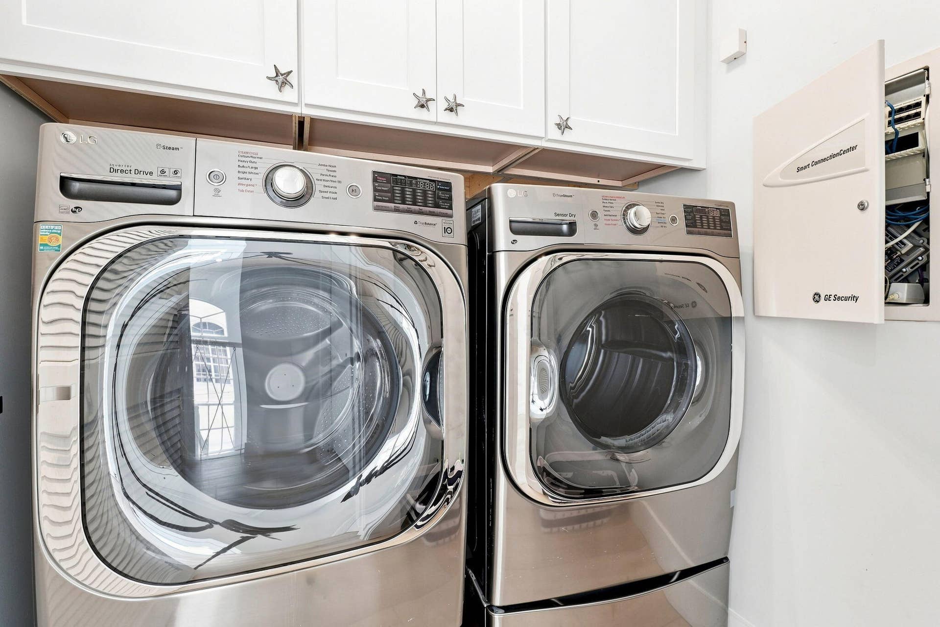 White cabinetry with decorative pulls, stainless steel washer and dryer units and a wall-mounted control panel