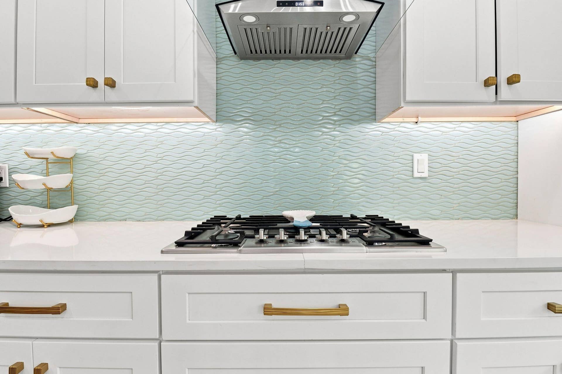 White shaker cabinetry with gold-tone hardware, built-in cooktop, stainless steel range hood, light-toned countertop and a wave-patterned tile backsplash