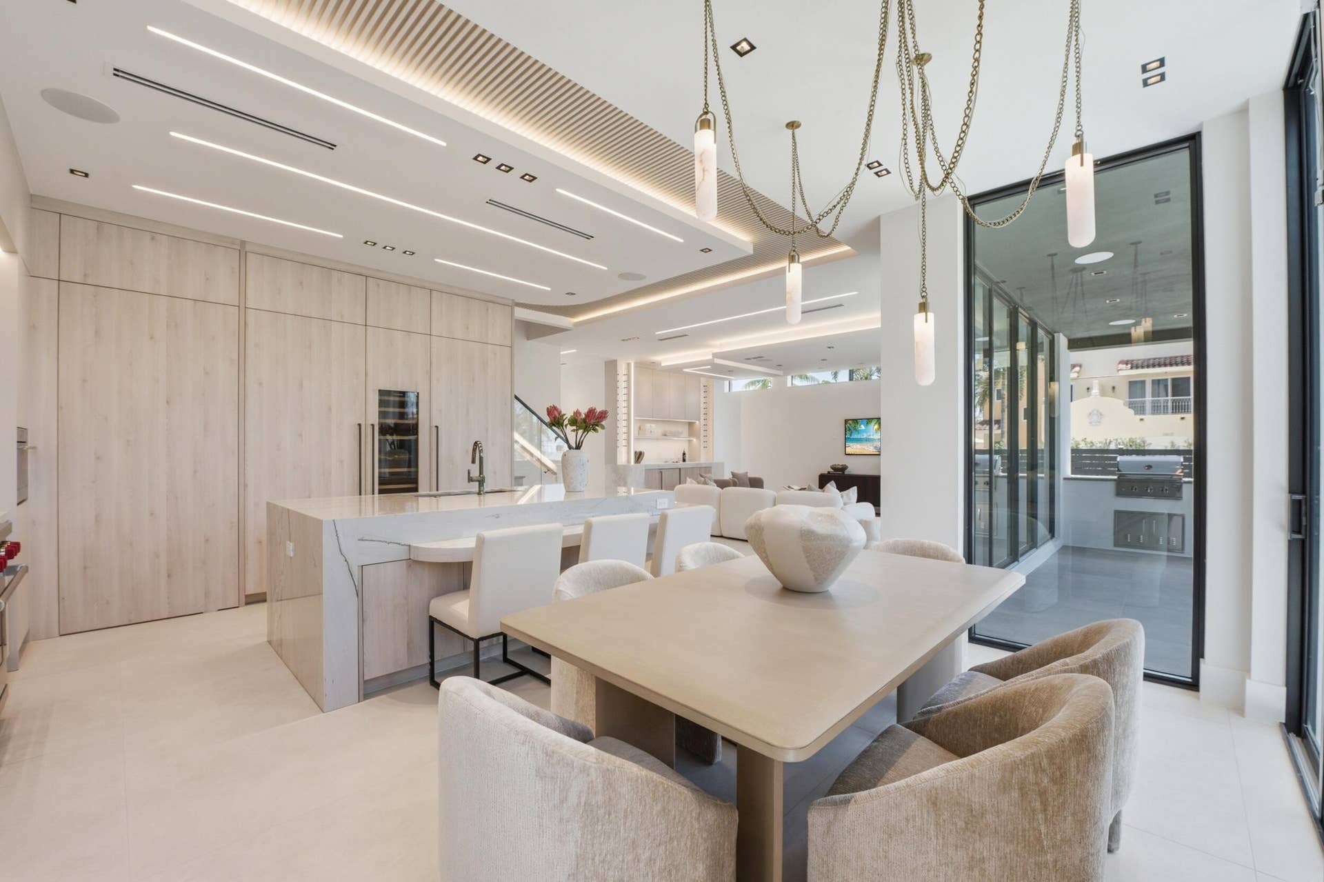 Custom kitchen cabinetry with integrated paneled appliances, large center island with waterfall countertop, recessed and linear ceiling lighting, and a multi-pendant dining chandelier