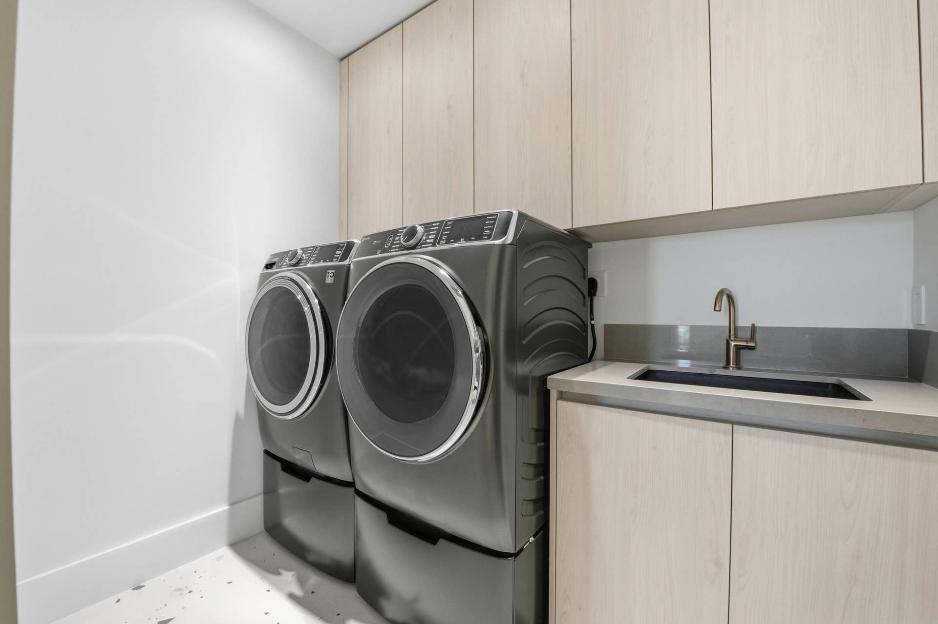 Integrated laundry area featuring light wood-finish cabinetry, a single basin sink with a bronze-tone faucet, and a grey countertop