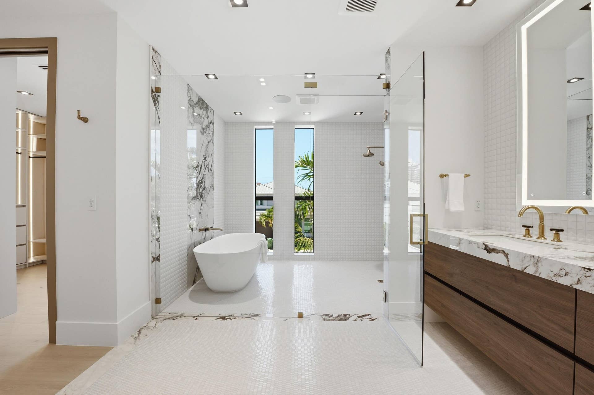 Freestanding soaking tub, glass-enclosed shower, dual vanity with wood-finish cabinetry, marble-look countertops, and gold-tone fixtures