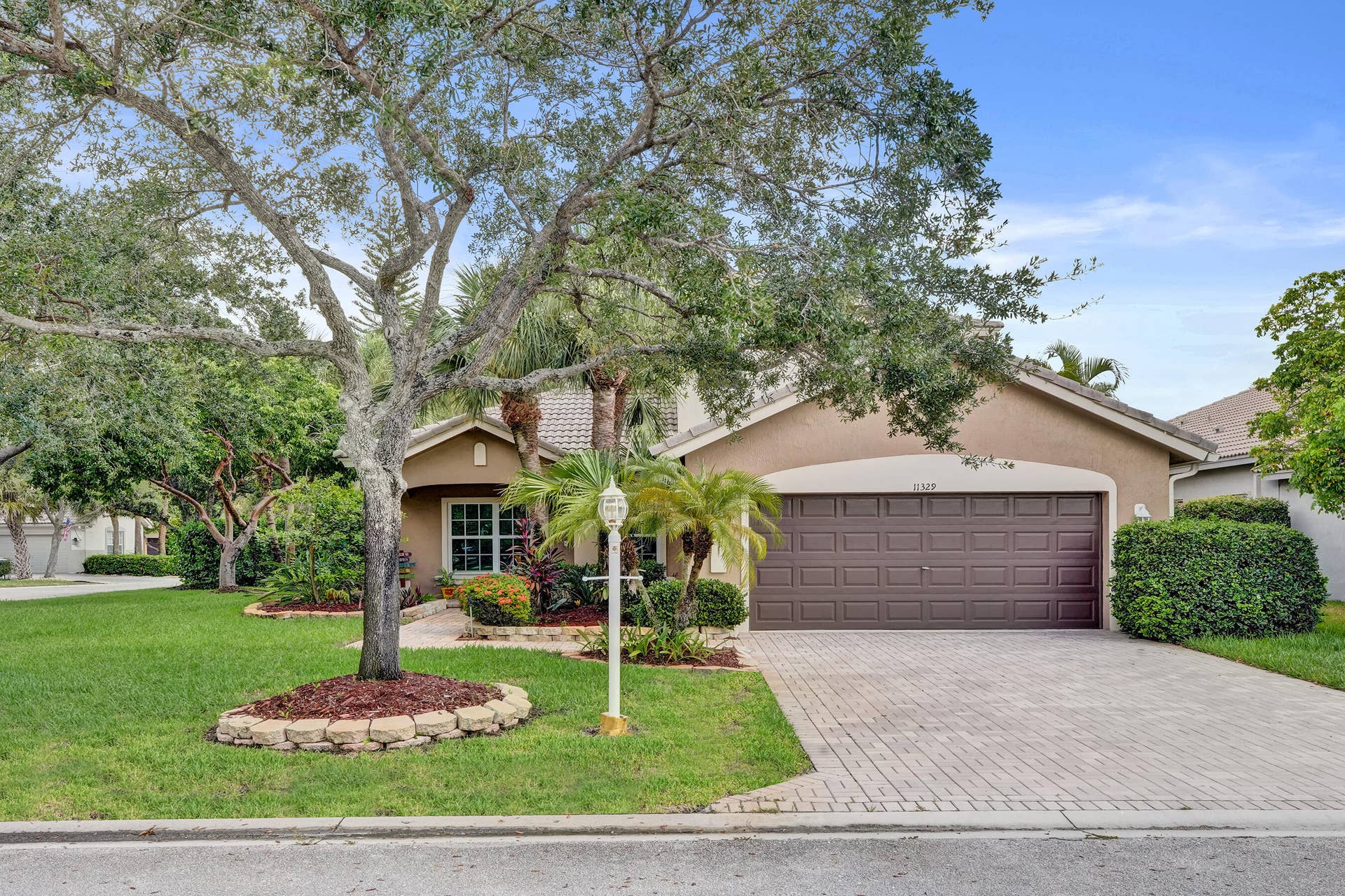 Welcoming curb appeal featuring a mature shade tree with circular mulch bed, manicured lawn, and professional hardscape paver driveway