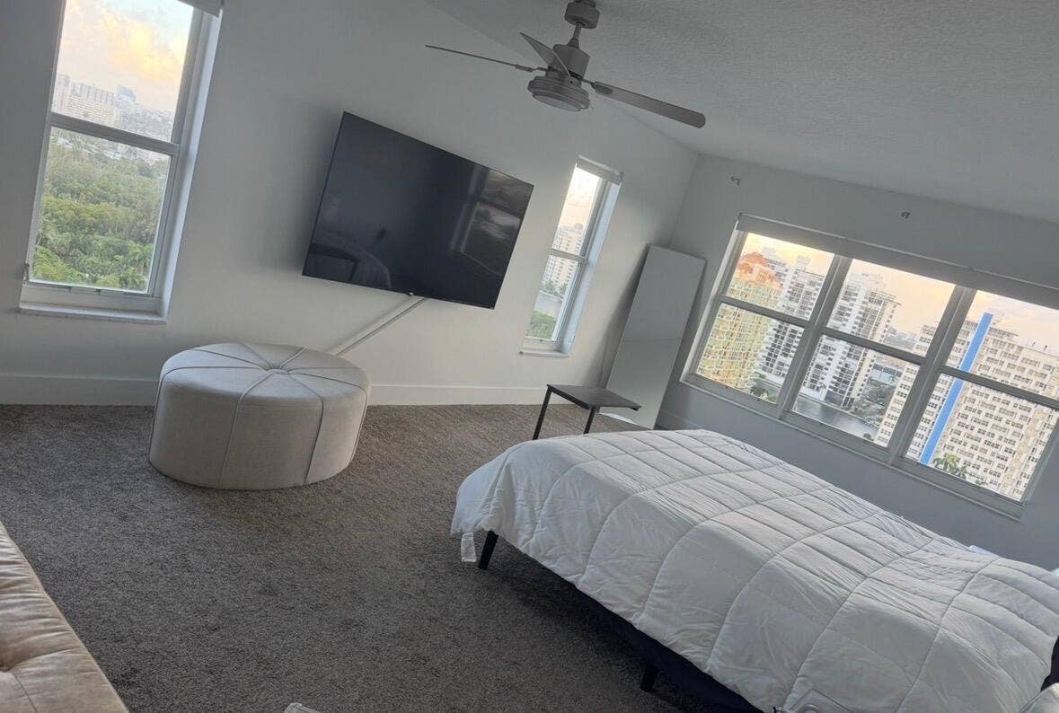 Wall-mounted television with visible wiring conduit, neutral-toned carpet, white walls, and multiple windows providing cityscape and greenery views