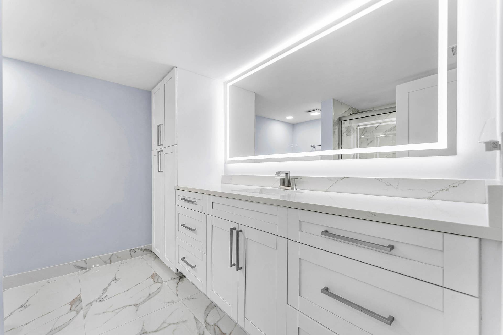White shaker cabinetry with brushed nickel hardware, tile flooring, illuminated mirror, and a white countertop with integrated sink