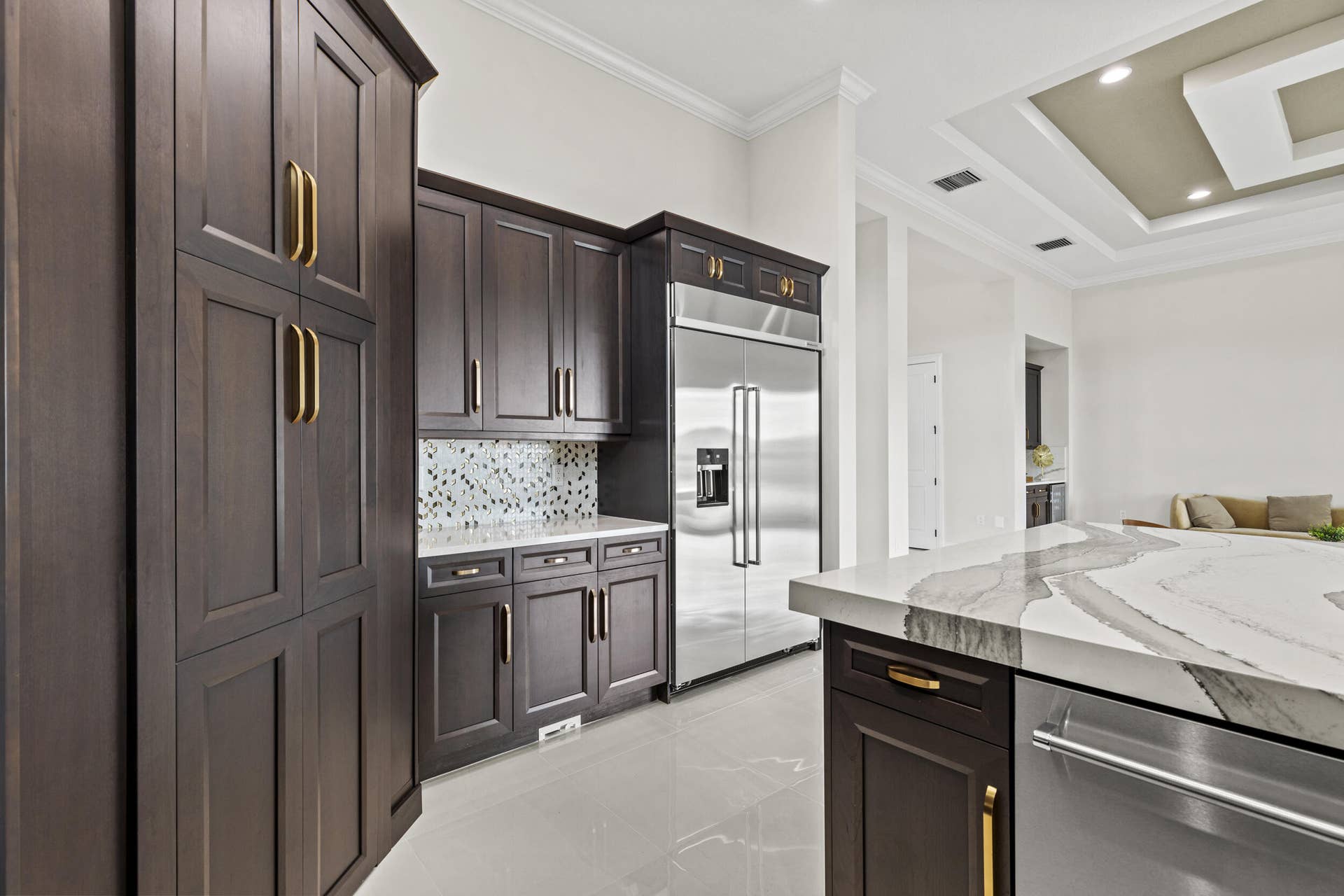 14976 Woodlot Court, Westlake, FL 33470 Dark wood cabinetry with gold-tone hardware, stainless steel refrigerator, patterned tile backsplash, light-toned flooring, and a tray ceiling