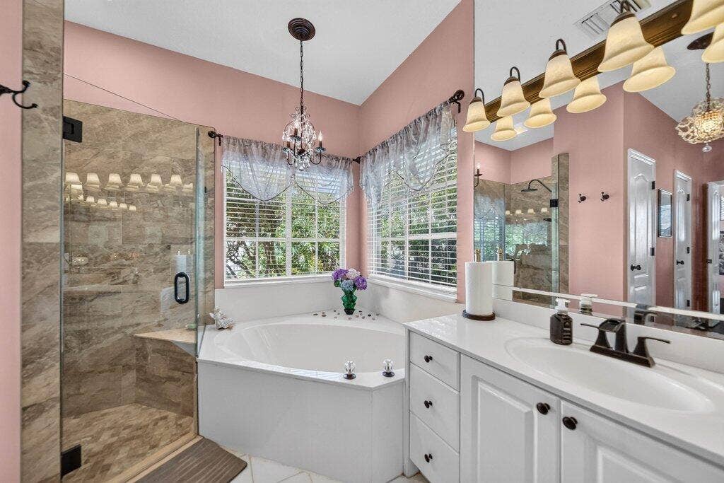 Tiled walk-in shower with a glass enclosure, corner soaking tub, dual-sink vanity with white cabinetry, and a chandelier fixture