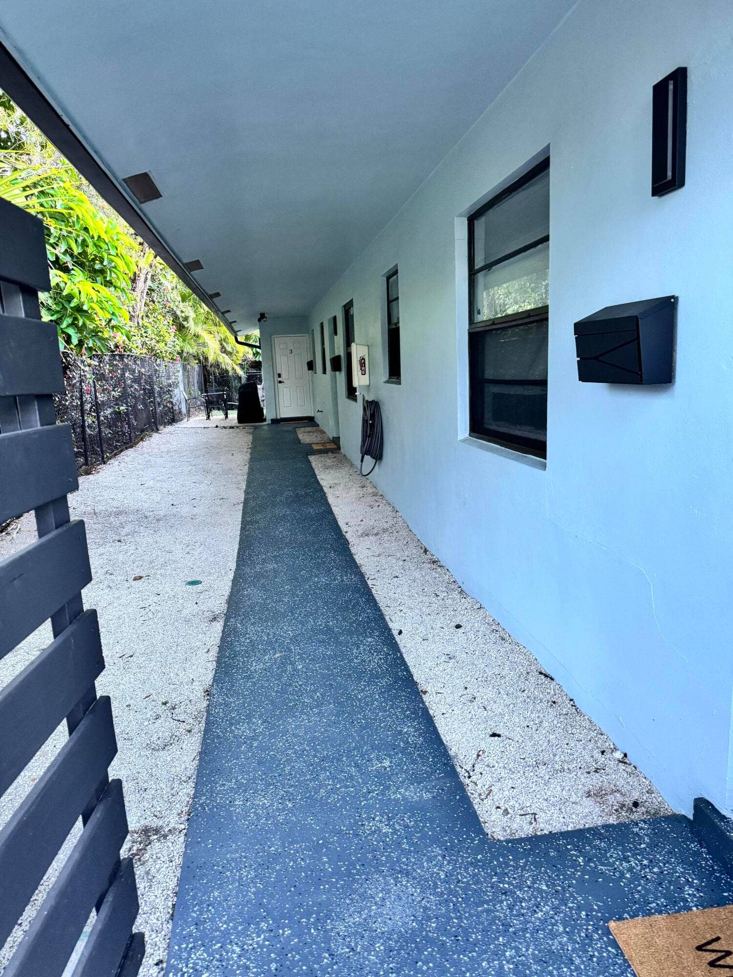 Covered walkway with a blue-painted concrete path, white stucco exterior, black-framed windows, and a modern black mailbox
