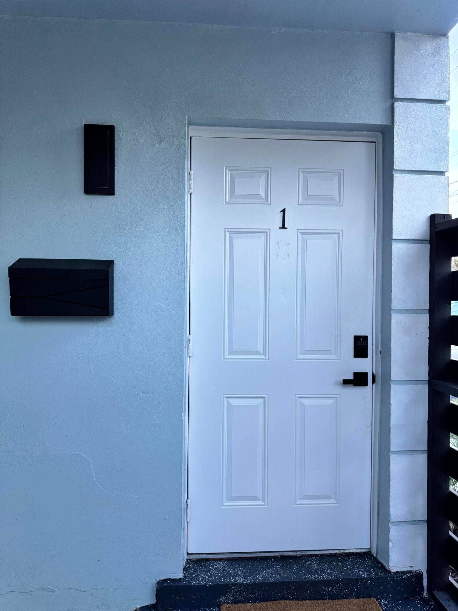 Panel door with black hardware, flanked by a black wall sconce and black mailbox