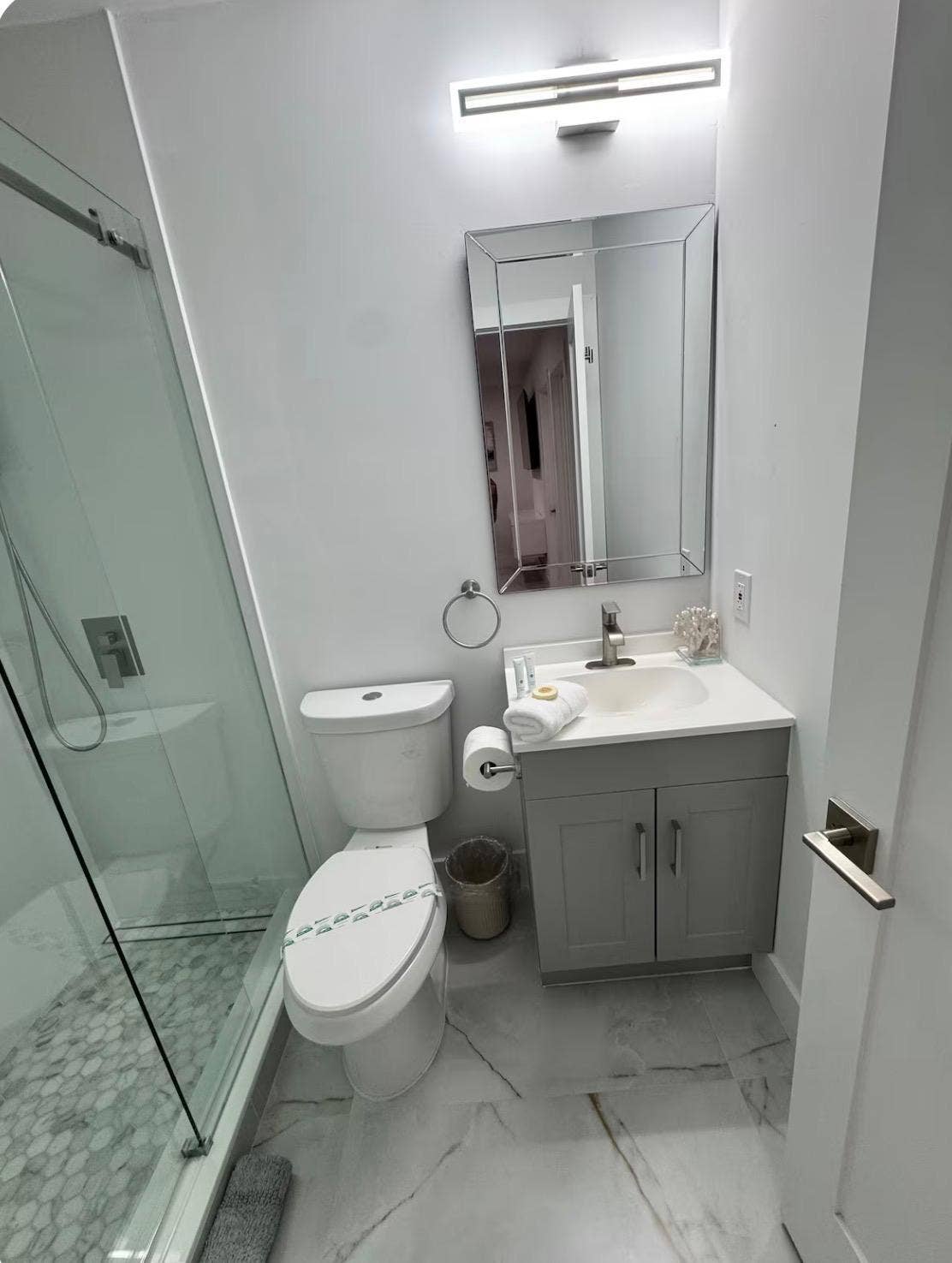 Glass-enclosed shower with pebble-style flooring, white vanity with gray cabinetry, rectangular mirror, and contemporary linear lighting fixture