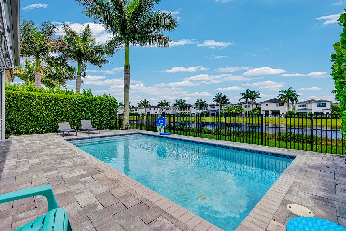 9046 CHAUVET Way, Boca Raton, FL 33496 Rectangular swimming pool with integrated steps, surrounded by paver patio surfacing, mature palm trees, and a privacy hedge