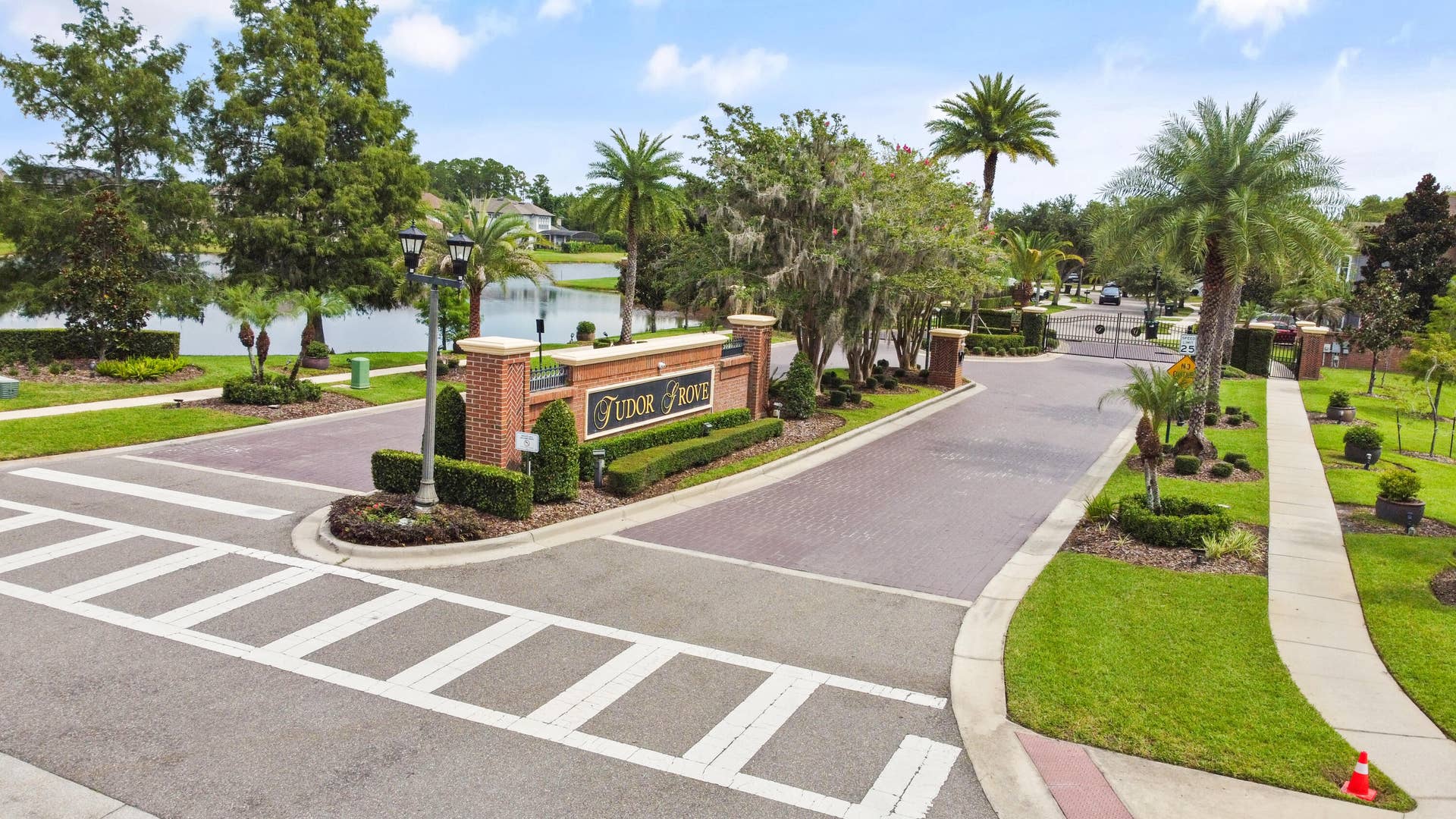 16437 TUDOR GROVE Drive, Orlando, FL 32828 Brick community entrance monument with "Tudor Grove" signage, surrounded by mature palm trees and manicured landscaping, featuring paved roadways and sidewalks