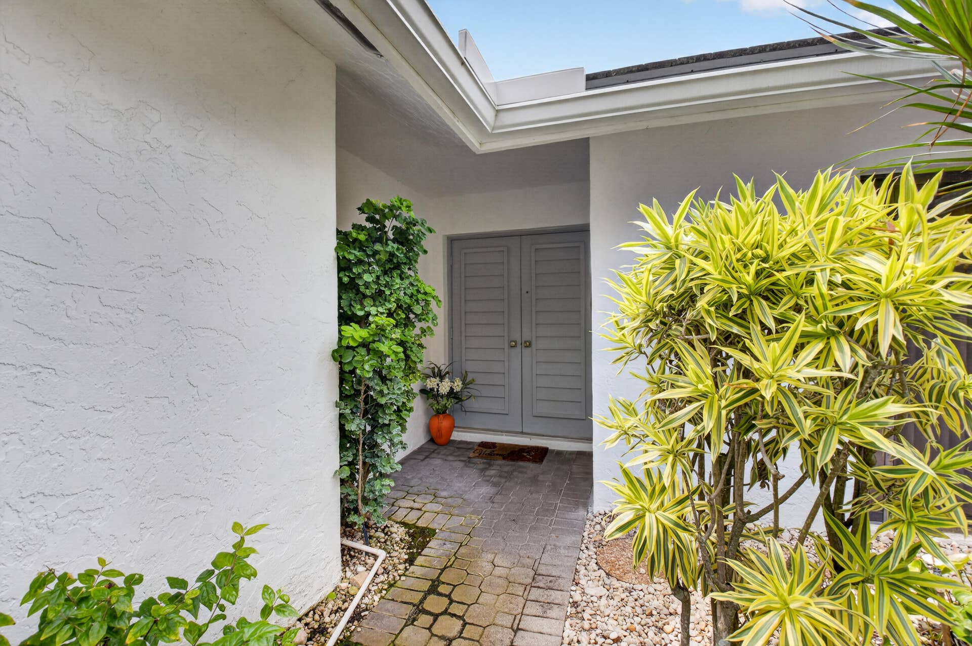 20065 Waters Edge Drive, #101, Boca Raton, FL 33434 Welcoming courtyard entry featuring crisp white brick exterior, dual gray panel doors, paved stone flooring, and lush tropical landscaping including vibrant golden foliage plants and climbing ivy accents