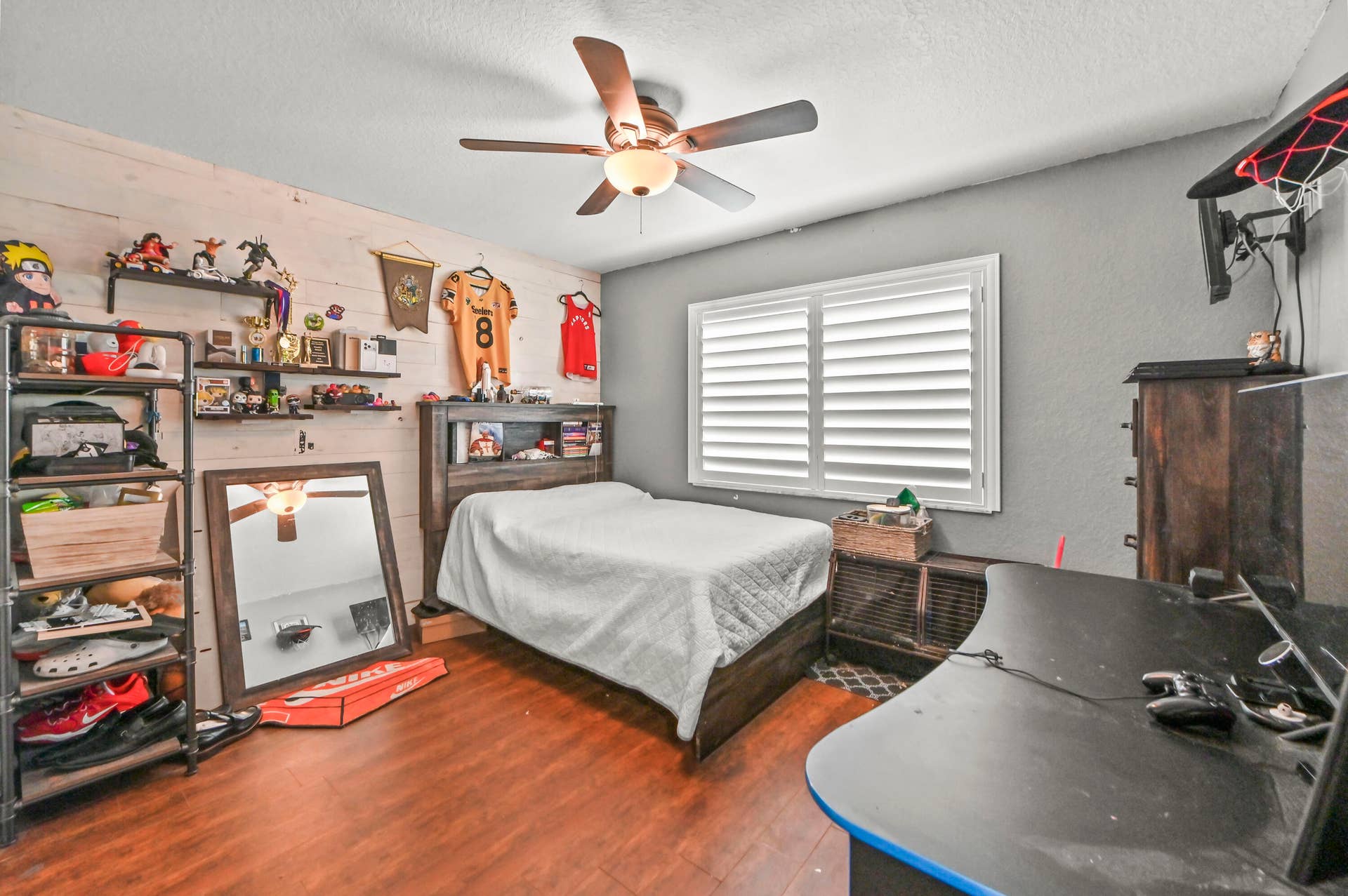 Wood-finish flooring, ceiling fan with integrated lighting, window with plantation shutters, accent wall with horizontal paneling