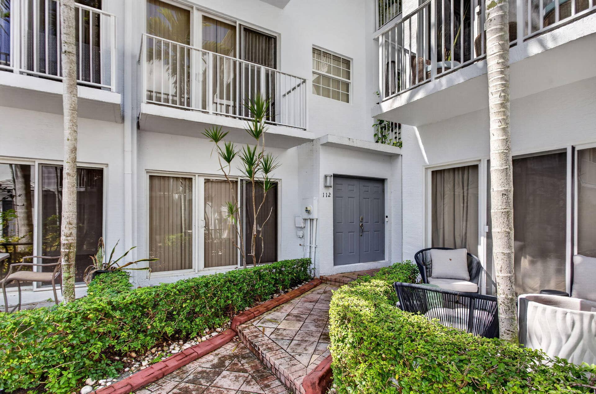 2725 NE 8TH Avenue, #112, Wilton Manors, FL 33334 White stucco exterior with a gray double entry door, multiple sliding glass doors, upper-level balconies with white railings, and a tiled walkway
