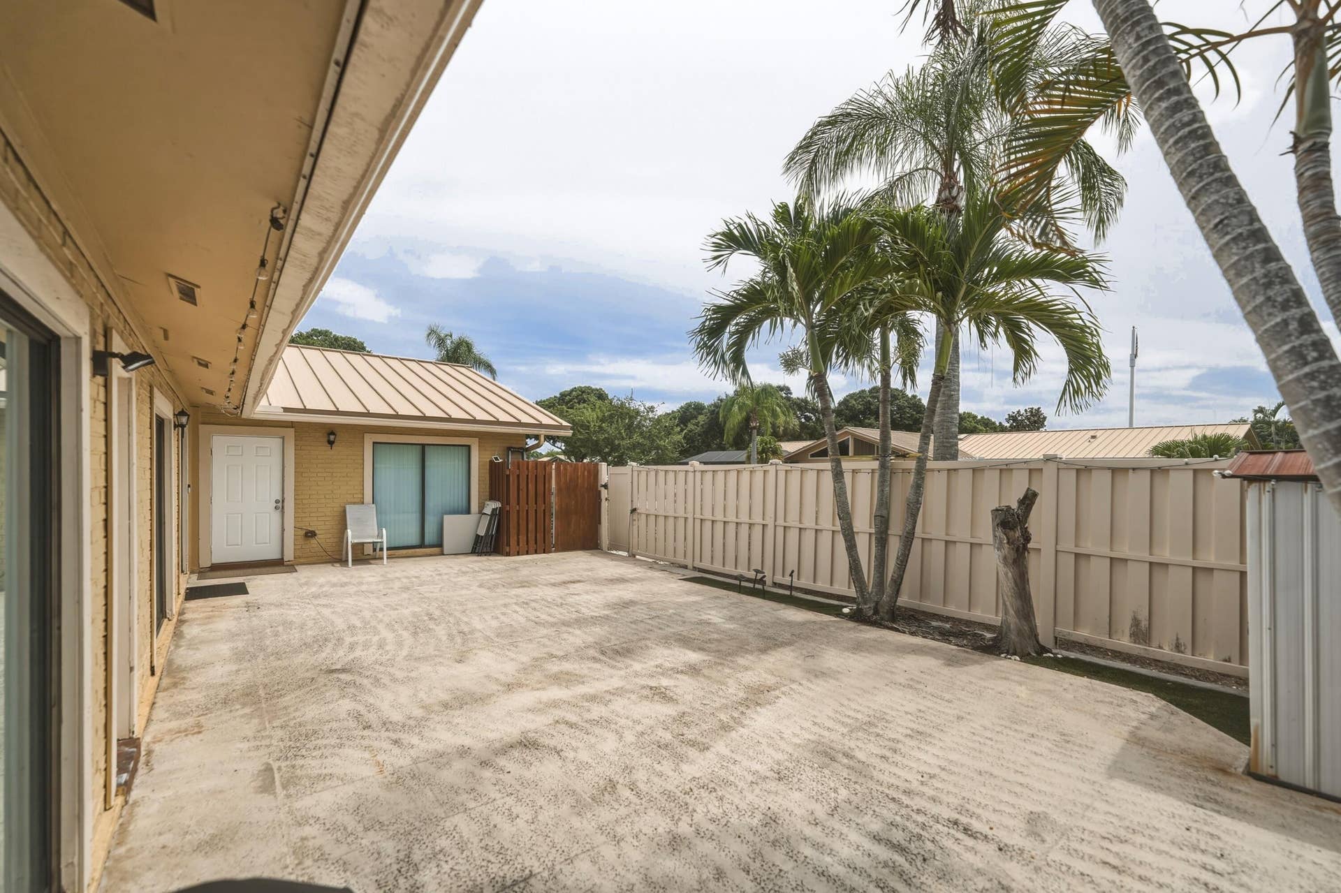 Expansive concrete patio with palm trees, a privacy fence, and a metal roof structure
