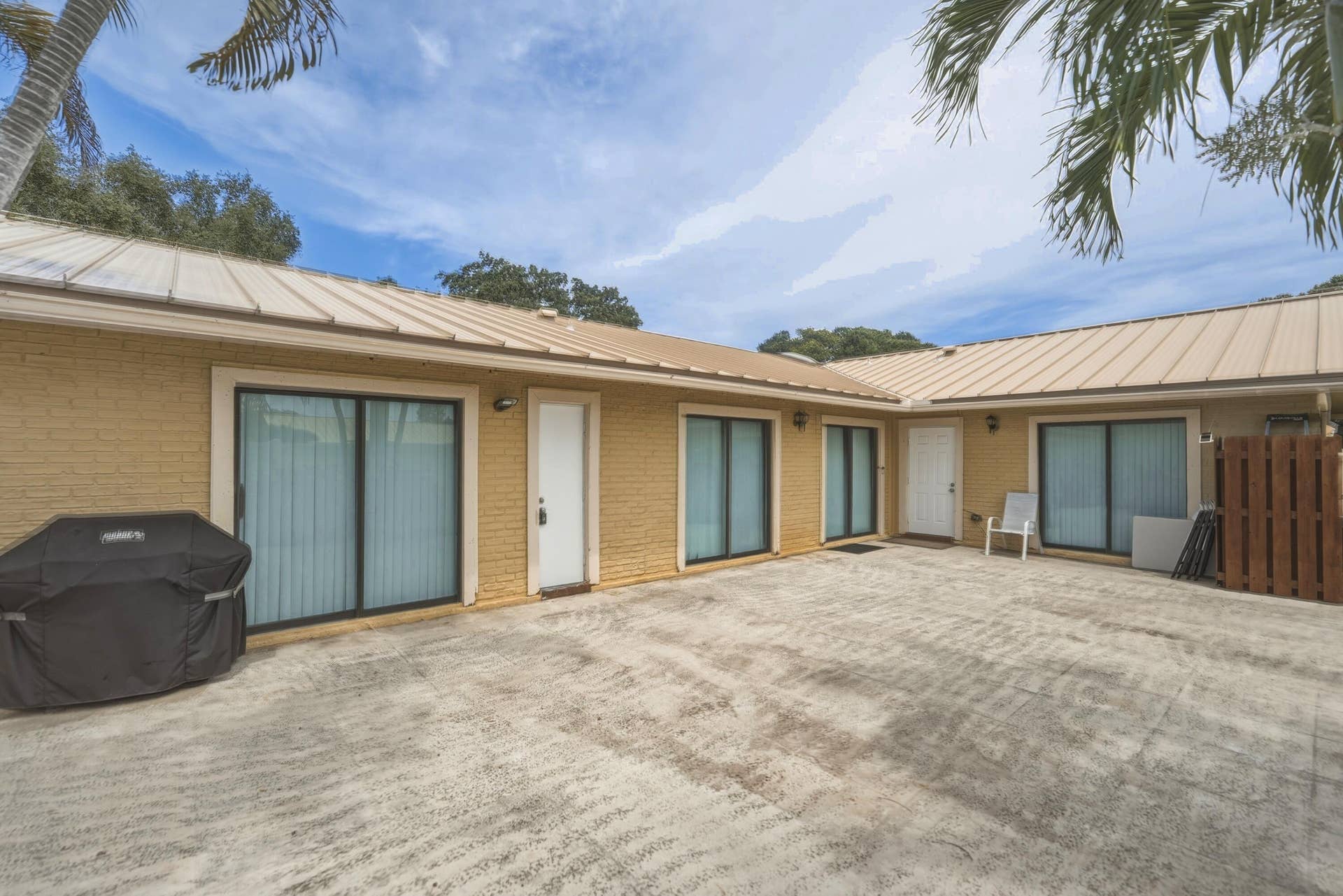 Expansive paved courtyard with multiple glass sliding door entries, brick veneer exterior, metal roof, and mature tree canopy providing natural shade