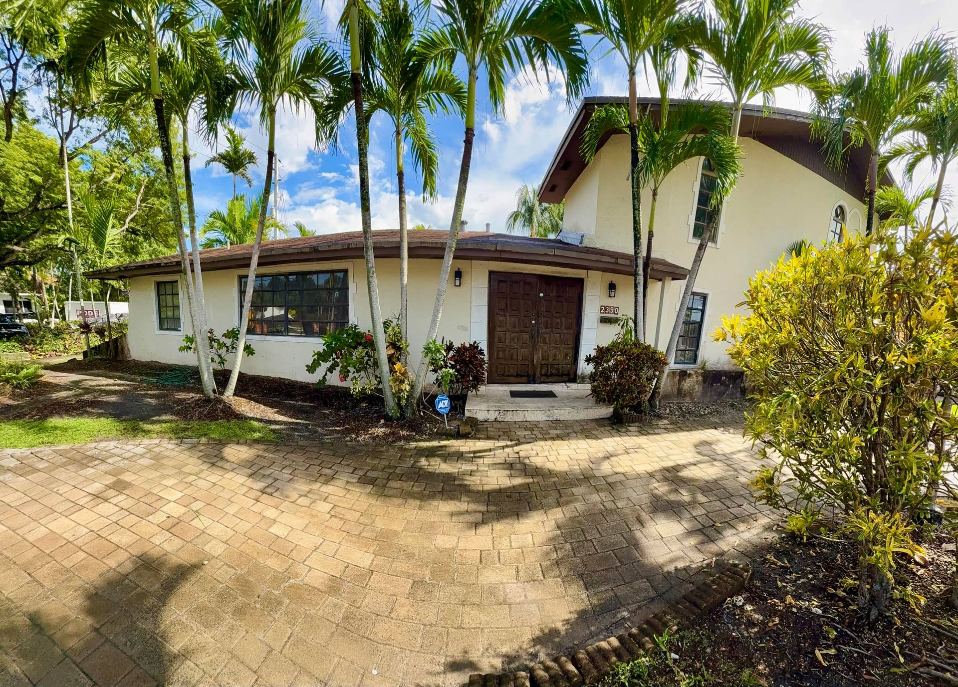 2390 NE 199th Street, Aventura, FL 33180 Stucco exterior with a brown roof and multiple palm trees, featuring a paved driveway and a wooden double-door entry