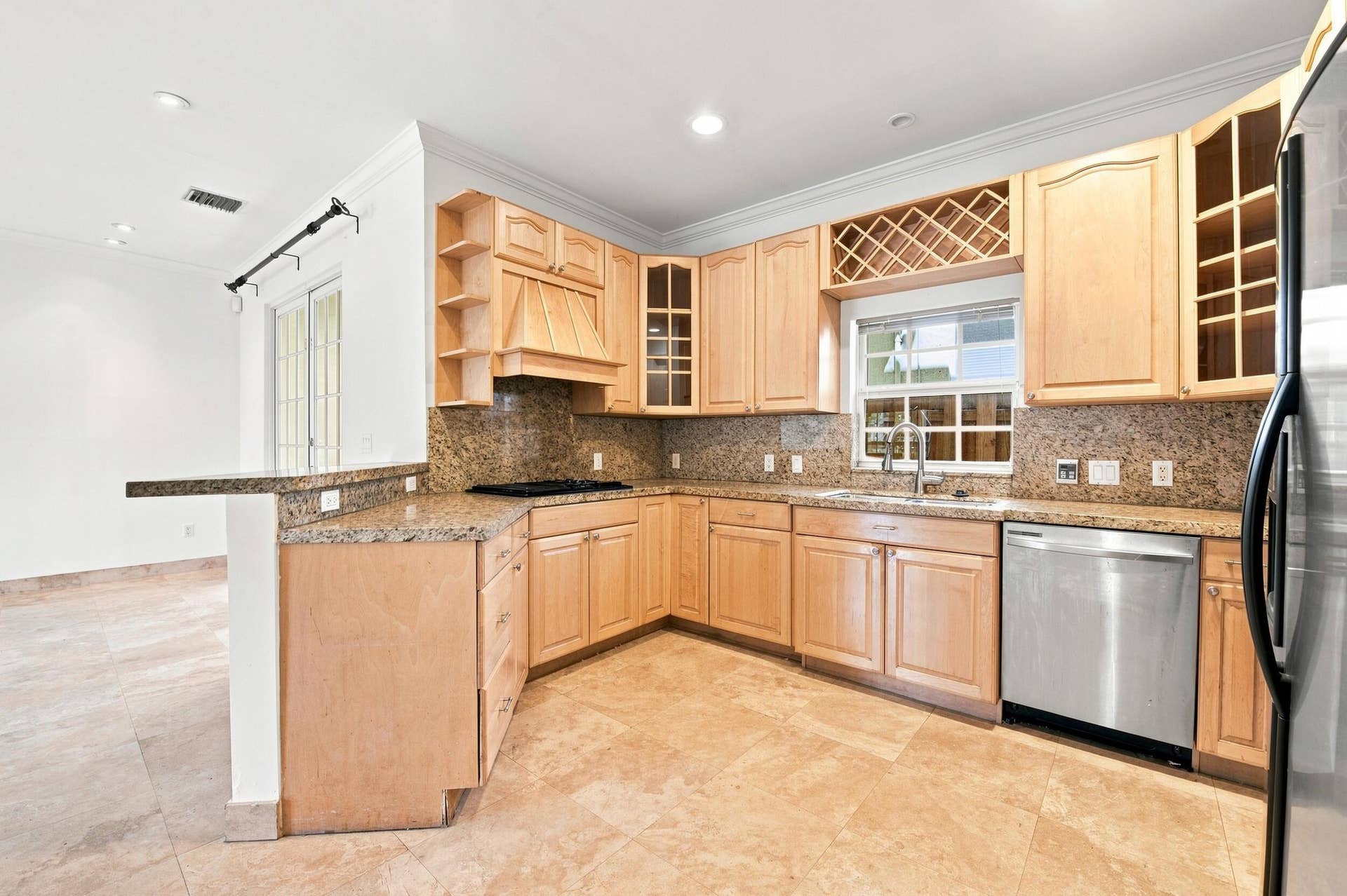 Spacious kitchen featuring warm wood-finish cabinetry with wine rack storage, granite countertops, stainless steel appliances including dishwasher, tile backsplash, and travertine flooring throughout the open layout