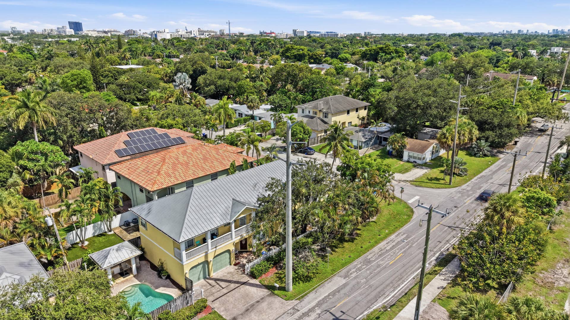 Modern residence featuring a pitched metal roof with solar panel installation, elevated on stilts with open ground-level foundation, complemented by a resort-style saltwater pool, mature landscaping with native palms and established trees, and situated on a corner lot with paved driveway access in an established neighborhood