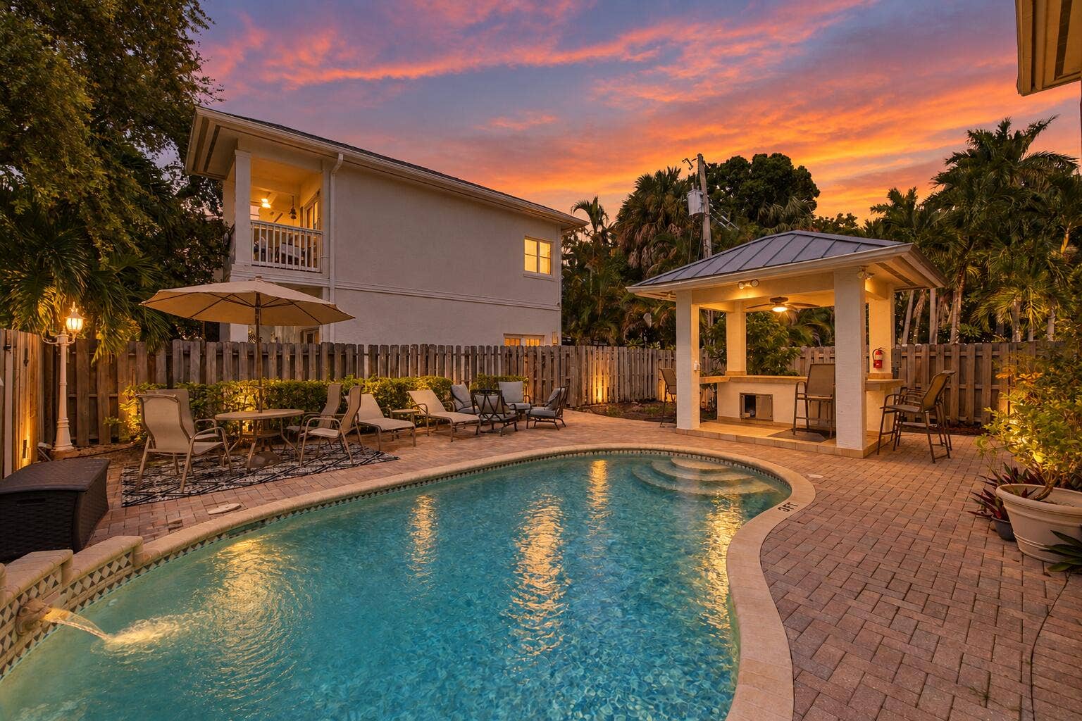Resort-style backyard oasis featuring a freeform saltwater pool with integrated lighting, expansive paver deck, covered loggia with ceiling fans, mature palm and tropical landscaping, and illuminated lounge seating areas