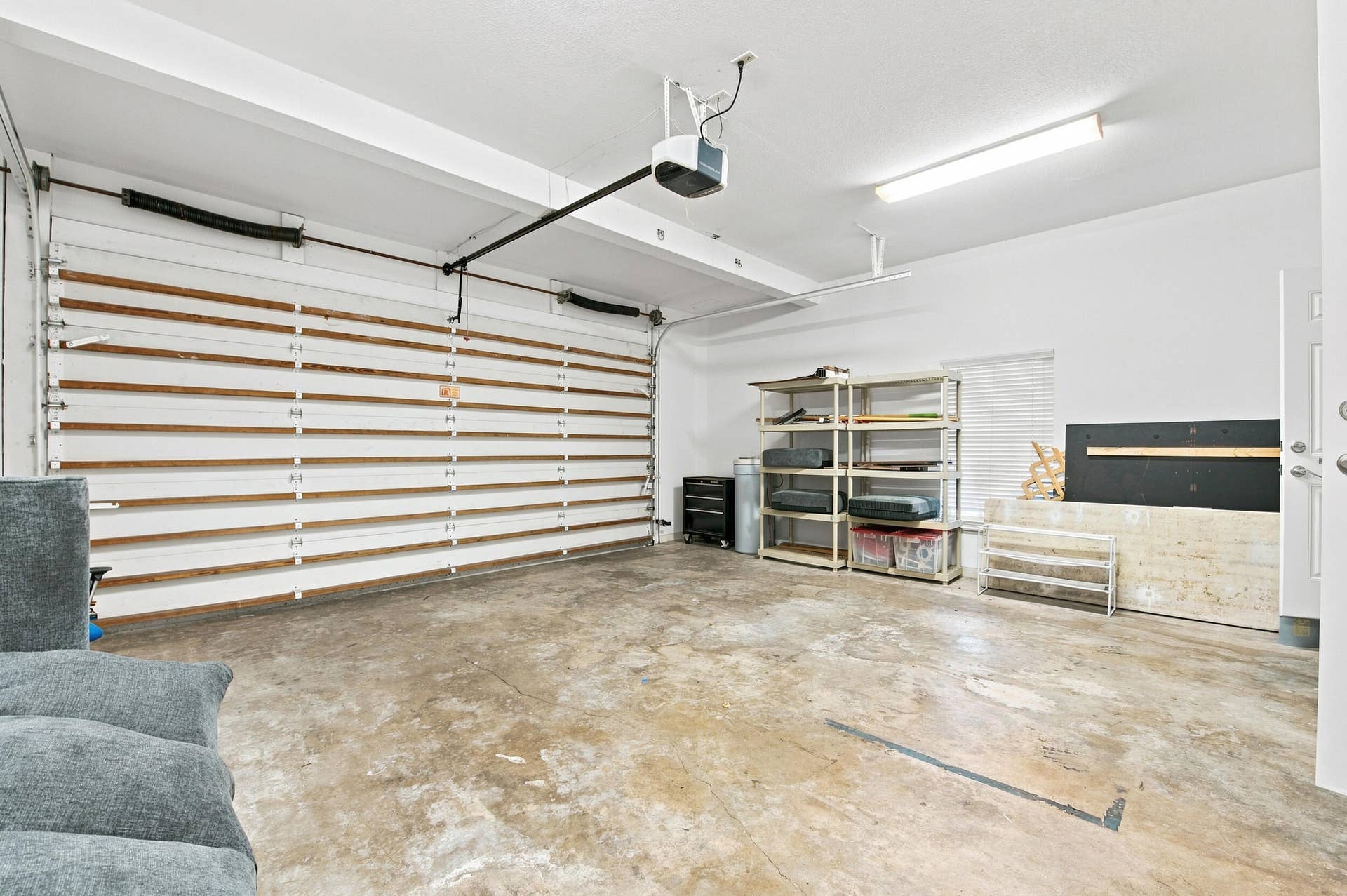 Spacious garage featuring epoxy-coated concrete flooring, overhead storage racks with wood-finish shelving, integrated ceiling-mounted lighting system, and white roll-up garage door with natural light entry