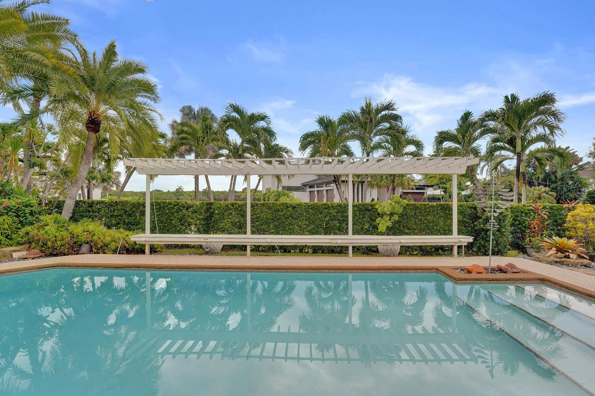 Swimming pool with integrated steps, surrounded by a hardscaping border, a white pergola, and lush perimeter landscaping