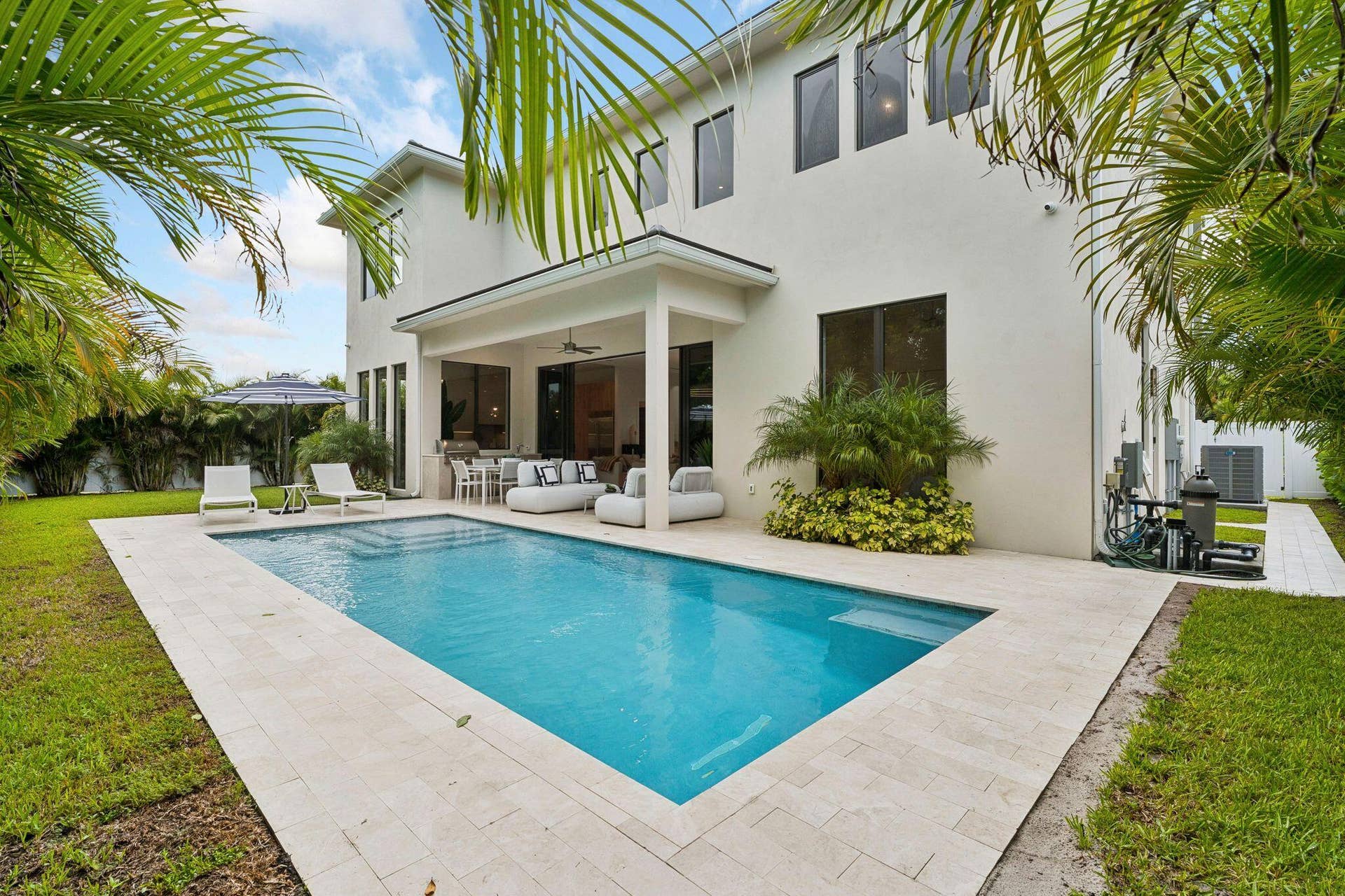 Spacious exterior featuring a rectangular pool, light-toned paver surround, covered patio with recessed lighting, and lush landscaping