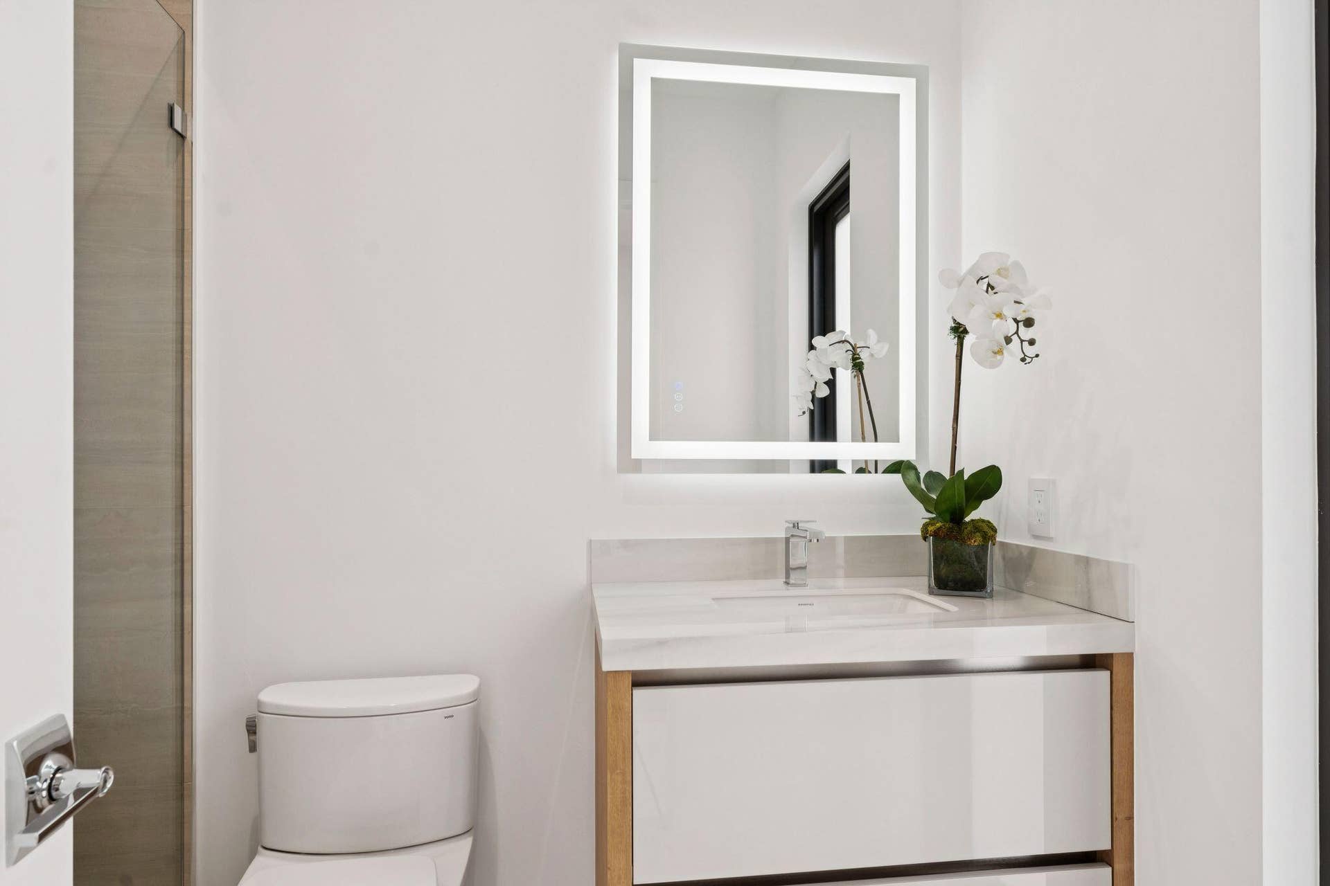 Backlit vanity mirror, single basin sink, white stone countertop, wood-finish vanity with white drawer fronts, and a modern toilet