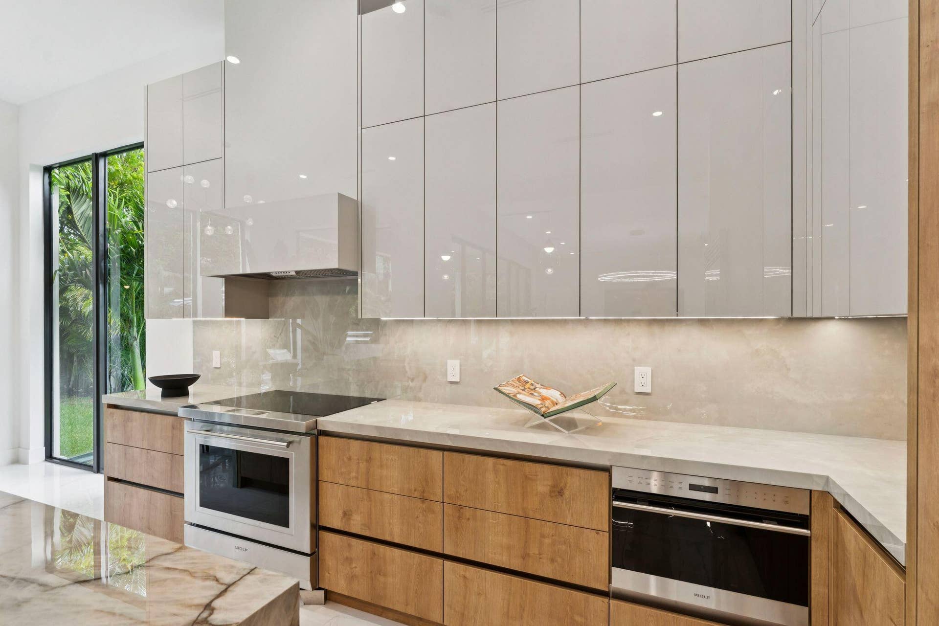 Sleek kitchen featuring light-toned upper cabinetry, wood-finish lower cabinetry, stone countertops, and integrated stainless steel appliances