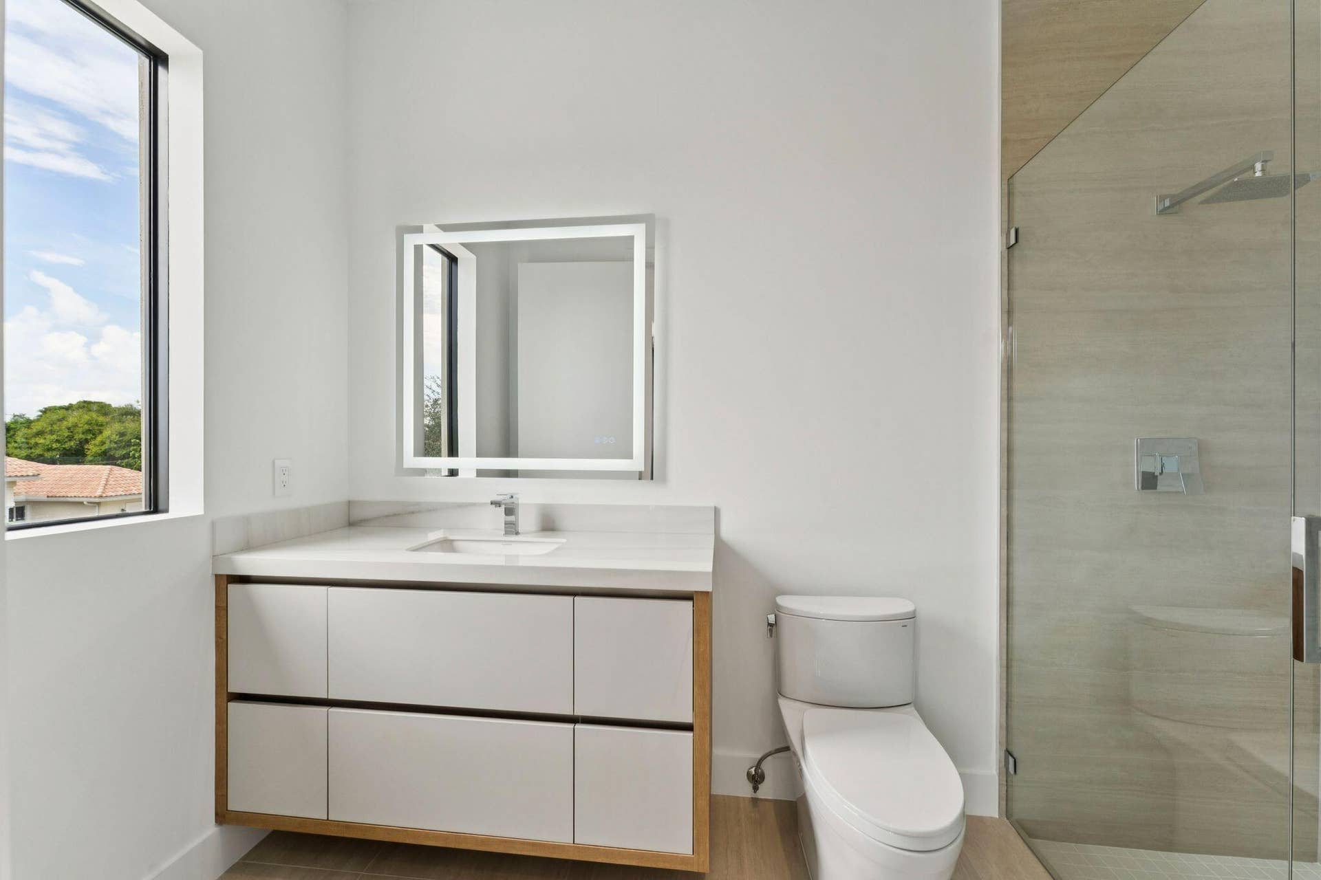 Modern primary bathroom featuring a floating vanity with white cabinetry and wood-finish base, quartz countertop, illuminated mirror with integrated lighting, chrome faucet, porcelain toilet, and a frameless glass-enclosed shower with tile surround and rainfall showerhead