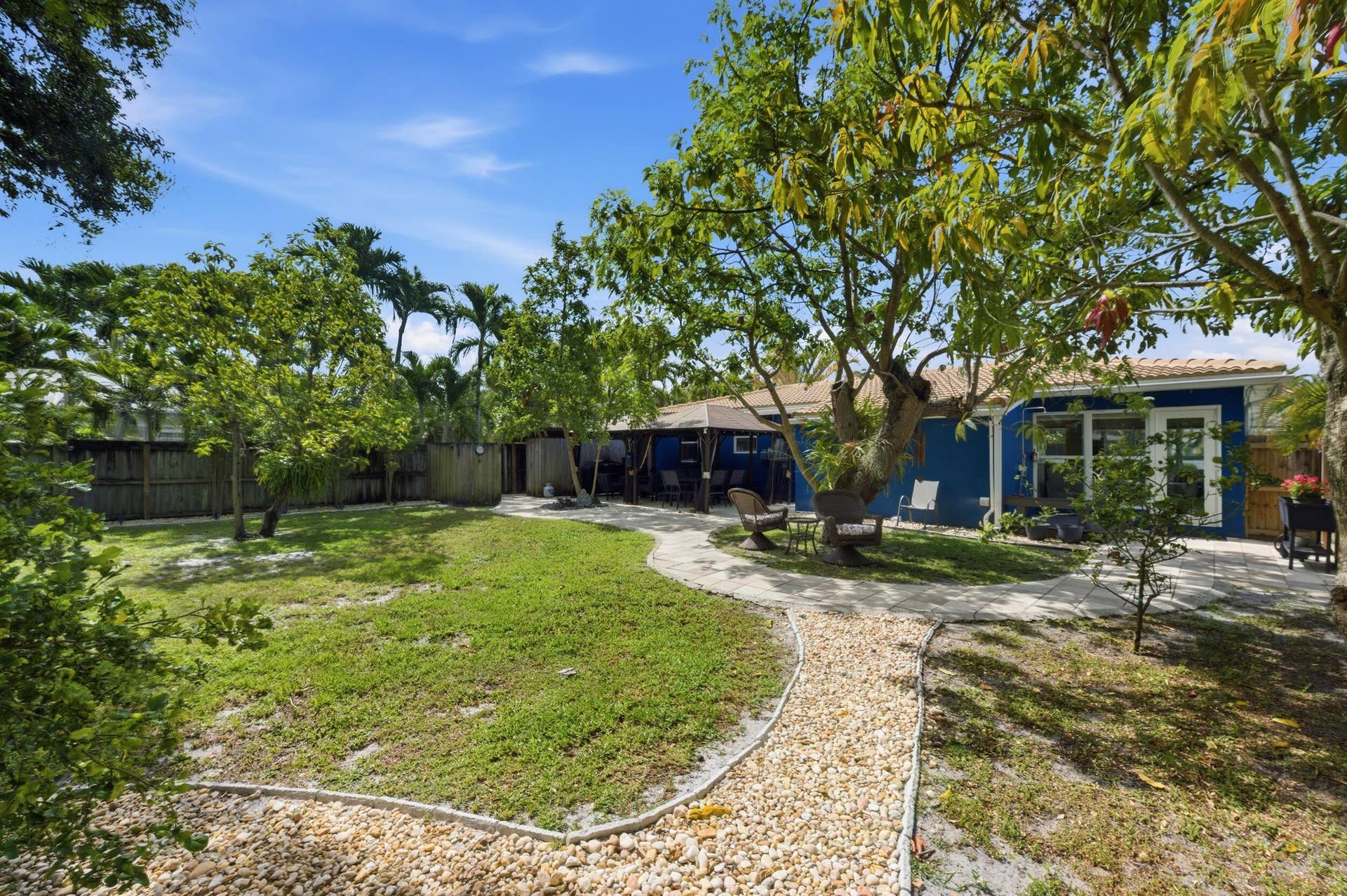 1421 SW 20 Street, Fort Lauderdale, FL 33315 Expansive backyard featuring a manicured lawn, established trees, a curved gravel pathway, a wooden privacy fence, and a blue exterior home with white framed windows