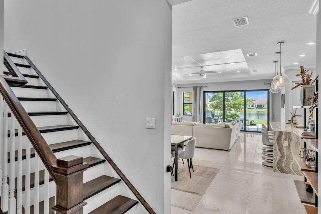 12143 S Baypoint Circle, Parkland, FL 33076 Modern interior featuring a wood-finish staircase with white risers and balusters, a spacious open-concept living area with large format tile flooring, sliding glass doors to outdoor spaces, a kitchen island with a waterfall countertop, and contemporary pendant lighting