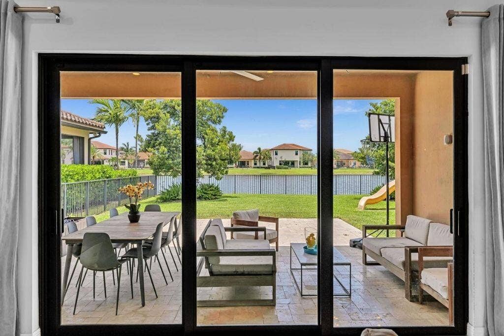 12143 S Baypoint Circle, Parkland, FL 33076 Sliding glass doors opening to a covered patio with tile flooring, featuring outdoor dining and lounge areas, and a manicured lawn overlooking a lake