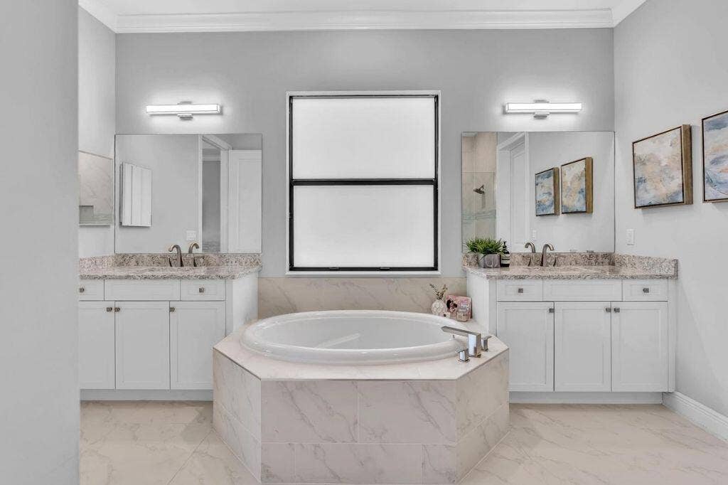 12143 S Baypoint Circle, Parkland, FL 33076 Soaking tub set in a tiled surround, flanked by two vanity cabinets with stone countertops and undermount sinks
