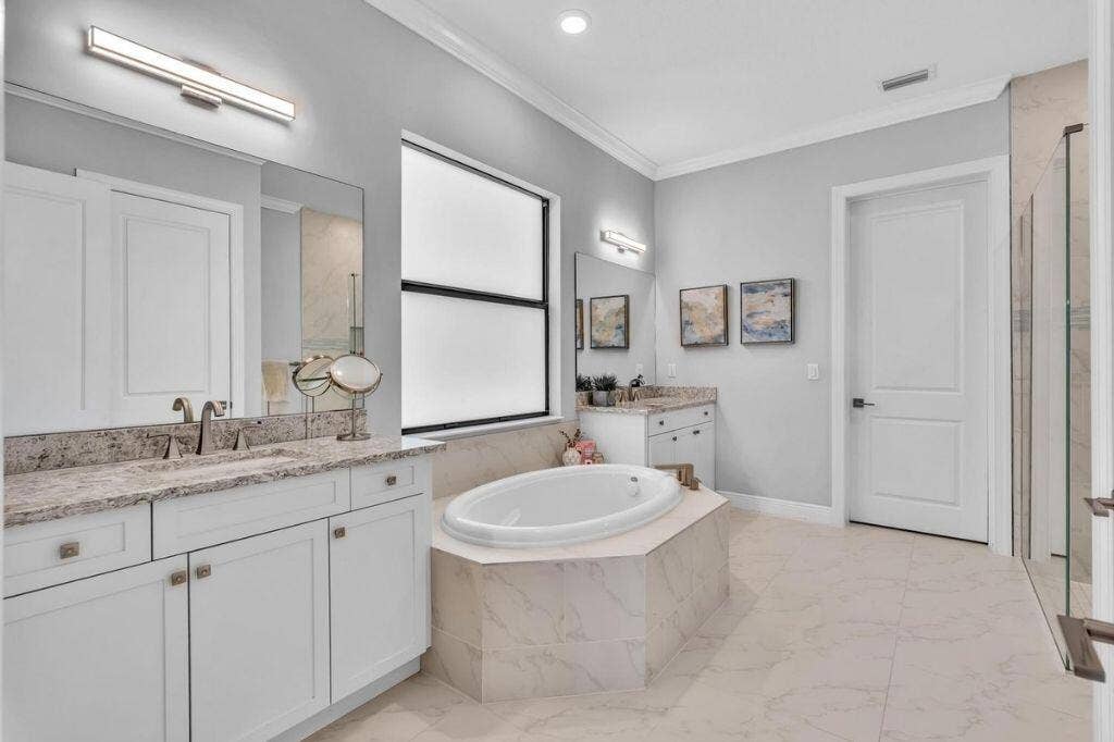 12143 S Baypoint Circle, Parkland, FL 33076 Light-filled bathroom featuring dual vanity stations with integrated sinks and granite countertops, a built-in soaking tub, a frameless glass shower enclosure, and large format tile flooring