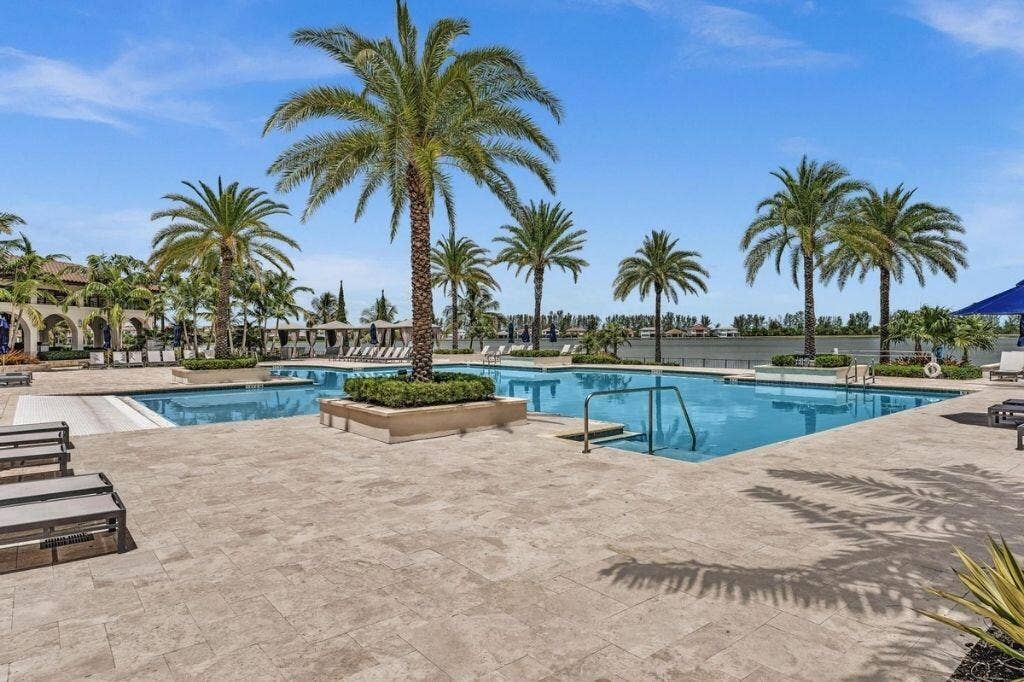12143 S Baypoint Circle, Parkland, FL 33076 Expansive pool area featuring multiple swimming pools, a stone paver deck, lush landscaping with palm trees, and a waterfront backdrop
