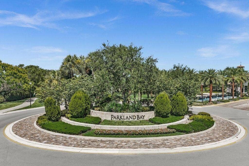 12143 S Baypoint Circle, Parkland, FL 33076 Roundabout entrance featuring a stone-clad "Parkland Bay" sign, lush landscaping, brick paver accents, and mature palm trees