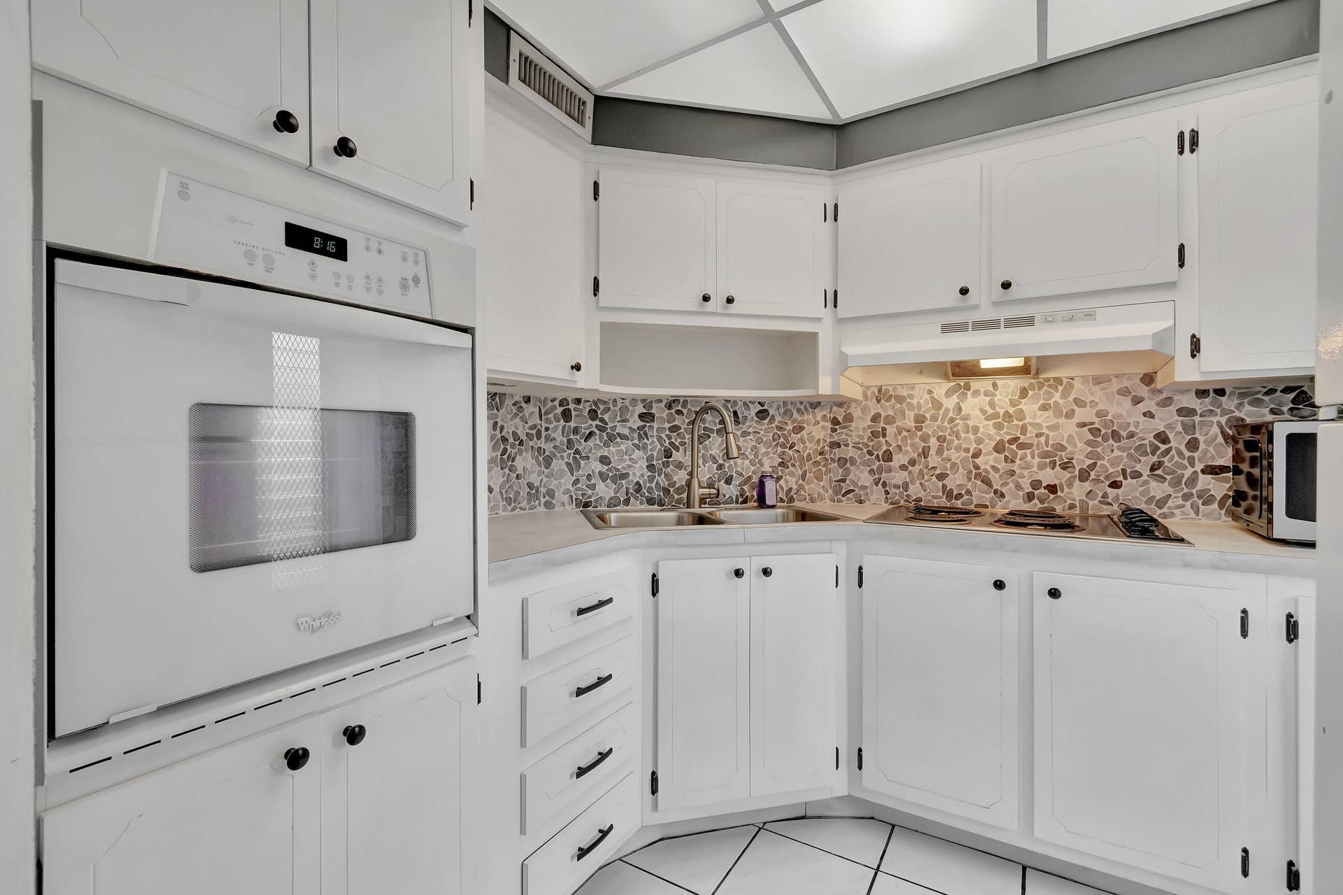 2161 NE 42nd Court, #116, Lighthouse Point, FL 33064 White cabinetry with black hardware, built-in oven, integrated cooktop, pebble-style tile backsplash, and white tile flooring