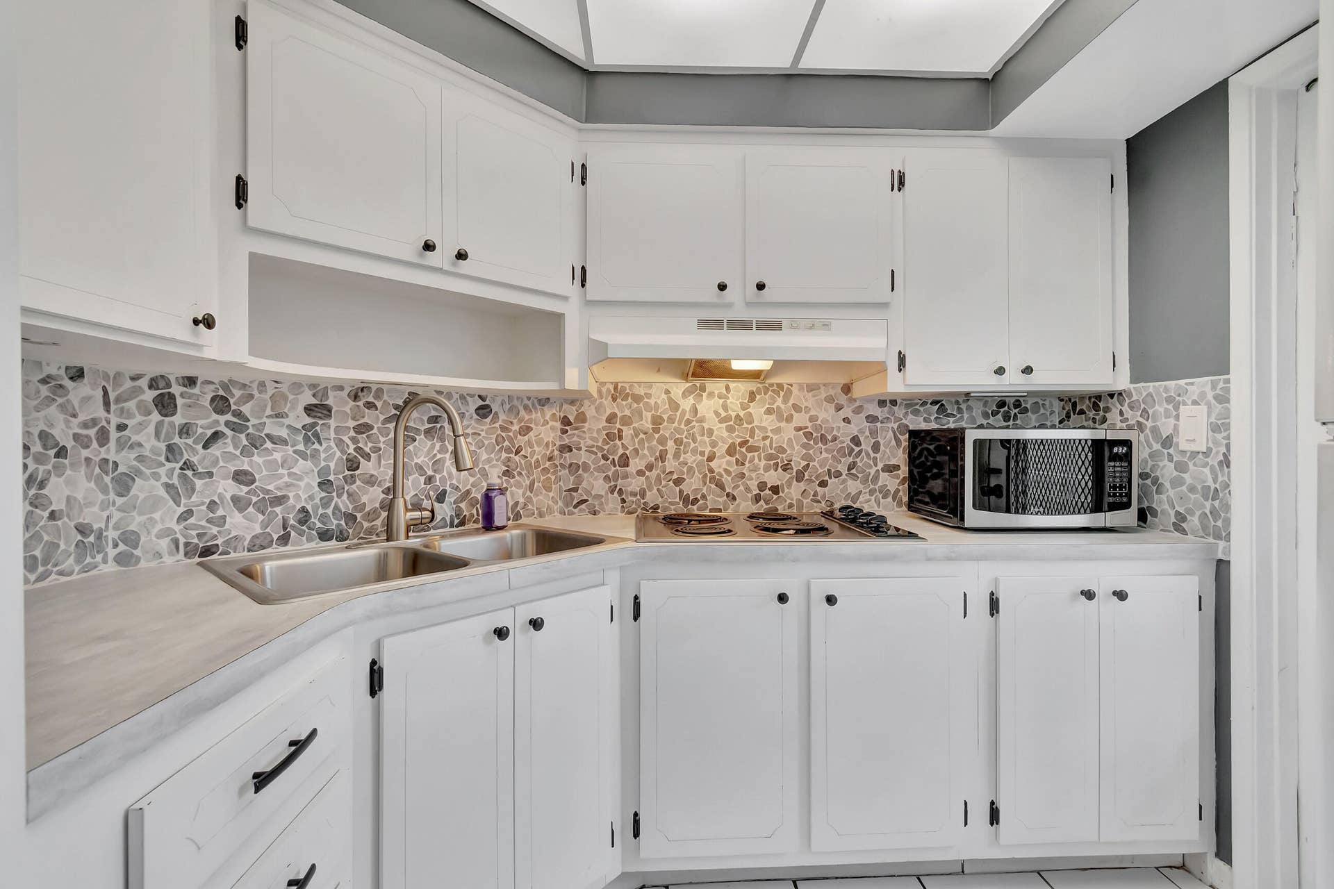 2161 NE 42nd Court, #116, Lighthouse Point, FL 33064 White shaker-style cabinetry with black hardware, stone tile backsplash, stainless steel double basin sink with gooseneck faucet, and light-toned countertops